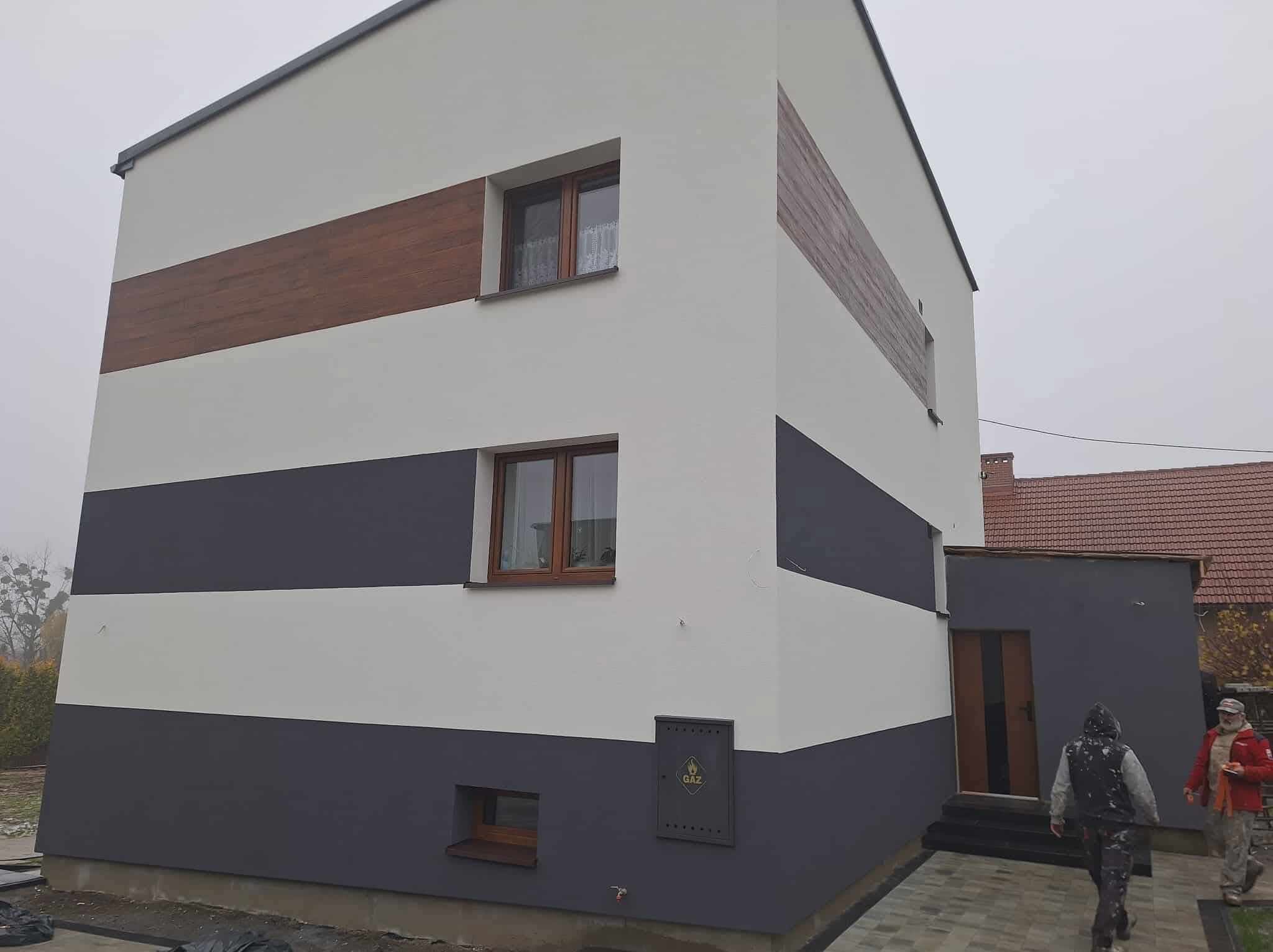 Dwukondygnacyjny dom po malowaniu elewacji w poziome pasy: szare, białe i imitujące drewno. Widoczna skrzynka gazowa i fragment chodnika z kostki.