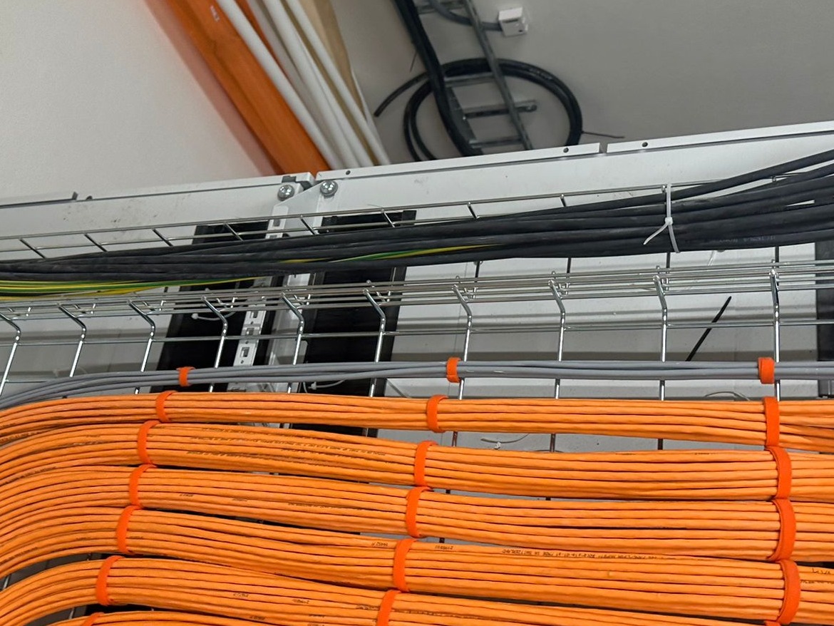Pomarańczowe i czarne kable ułożone w metalowej korycie pod sufitem, zabezpieczone pomarańczowymi opaskami. Widoczne inne kable w tle.