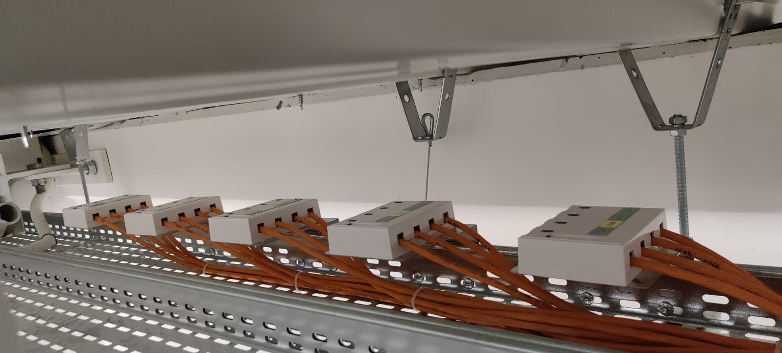 Rząd białych puszek łączeniowych z pomarańczowymi kablami, zamocowanych na perforowanej metalowej szynie pod sufitem. Widoczne elementy mocujące do sufitu.