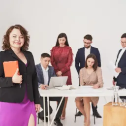 Uśmiechnięta kobieta w czarnej marynarce i fioletowej sukience z pomarańczowym notatnikiem na tle zespołu pracującego przy białym stole z laptopem i dokumentami.