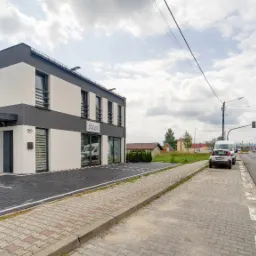 Dwukondygnacyjny budynek biurowy z szaro-białą elewacją, tablicą reklamową agencji nieruchomości 'Start' i adresem 99C, widok z chodnika z kostki brukowej w słoneczny dzień.