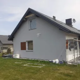 Dom jednorodzinny po tynkowaniu elewacji na szaro, z widocznymi śladami prac: rusztowanie, puste wiadra po farbie, rozłożone profile i taczka na trawniku.