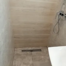 Wykończony brodzik kabiny prysznicowej z odpływem liniowym, wyłożony płytkami w odcieniach szarości i beżu, z widoczną baterią prysznicową i fragmentem umywalki.