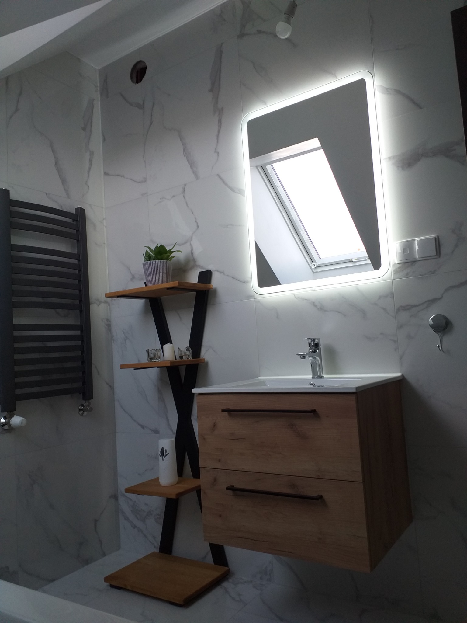 Nowoczesna łazienka z marmurowymi płytkami, podświetlanym lustrem, drewnianą szafką umywalkową i grzejnikiem drabinkowym. Widoczne okno dachowe odbijające się w lustrze, półki z dekoracjami...