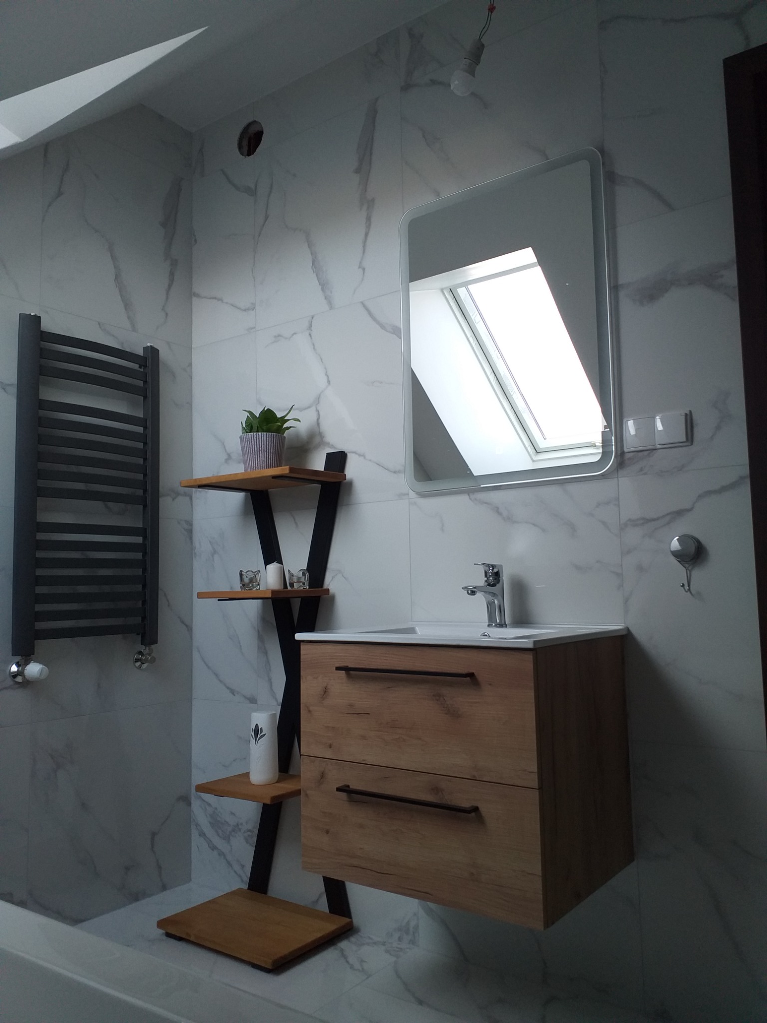 Nowoczesna łazienka z marmurowymi płytkami, drewnianą szafką pod umywalką, grzejnikiem drabinkowym i lustrem odbijającym okno dachowe.