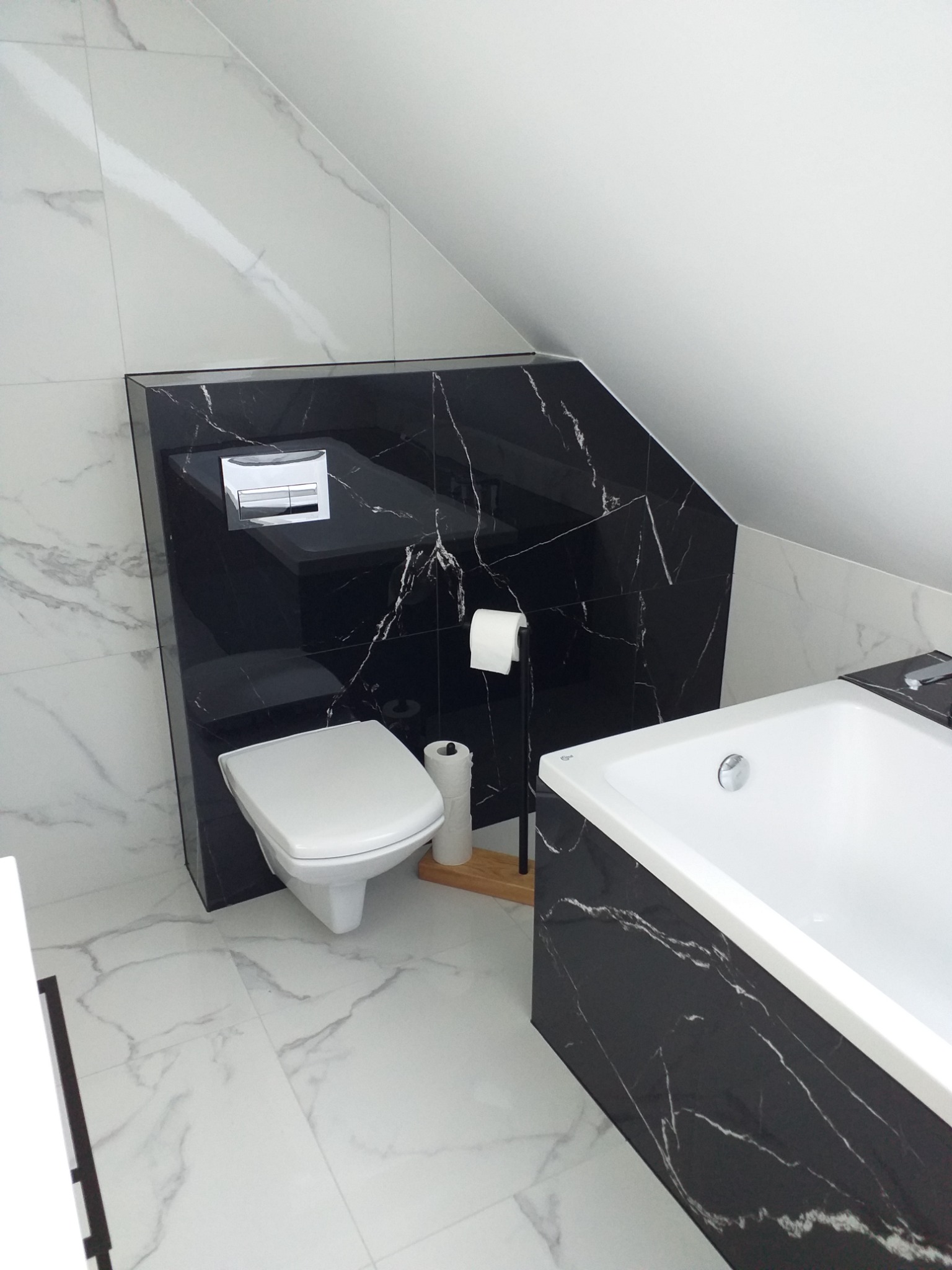 Nowoczesna łazienka na poddaszu z białymi marmurowymi płytkami na podłodze i ścianach, czarną marmurową zabudową WC i wanny.