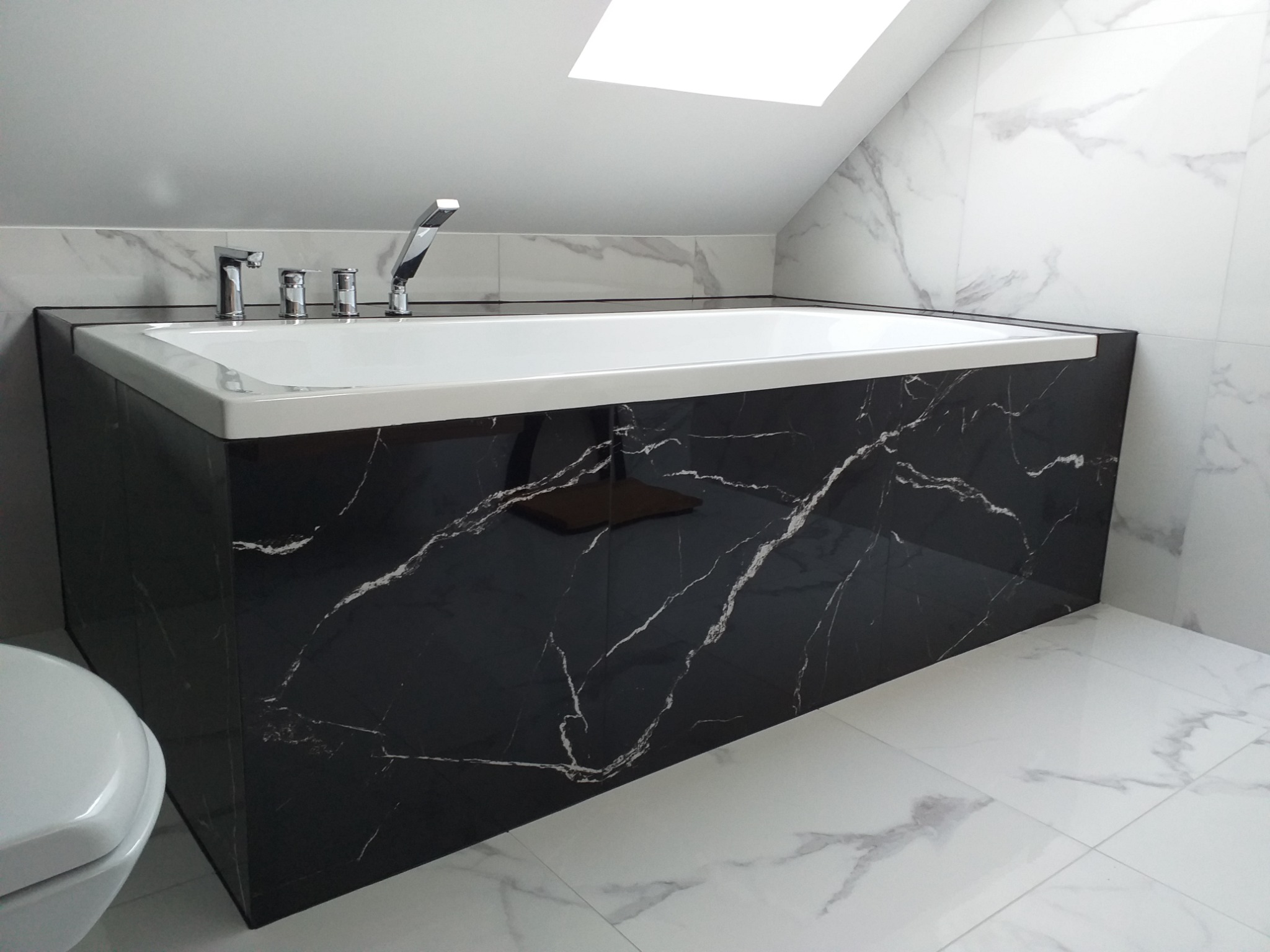 Nowoczesna łazienka na poddaszu z wanną obudowaną czarnym marmurem, baterią czterootworową i płytkami imitującymi biały marmur.