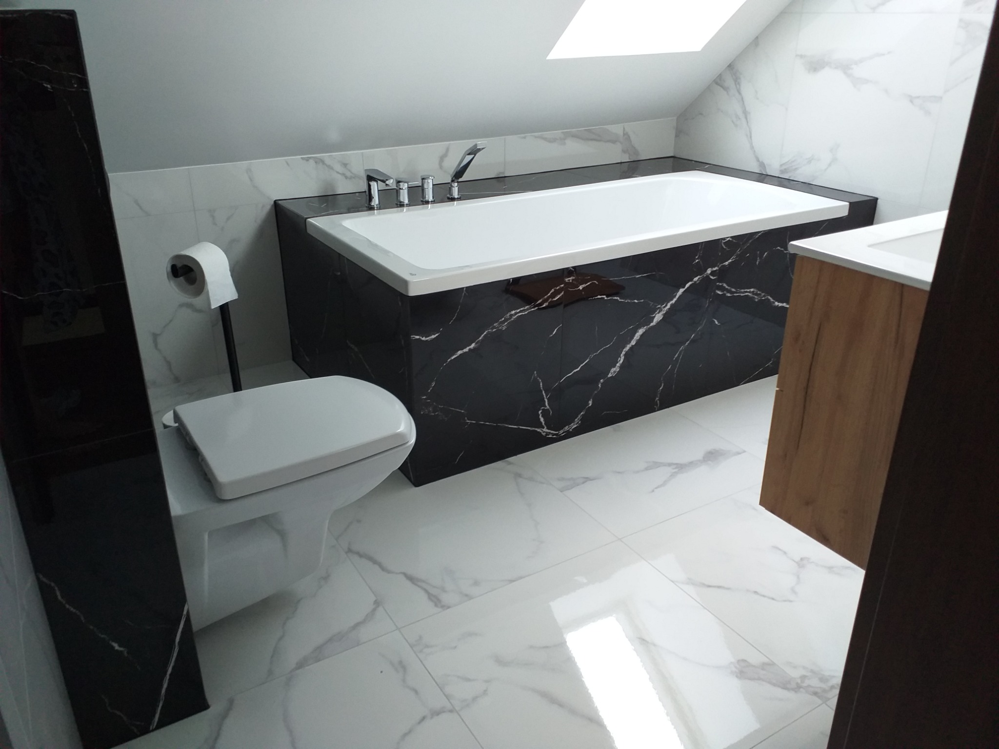 Nowoczesna łazienka na poddaszu z białą armaturą, wanną obudowaną czarnym marmurem, toaletą i umywalką z drewnianą szafką, wykończona białymi płytkami imitującymi marmur.