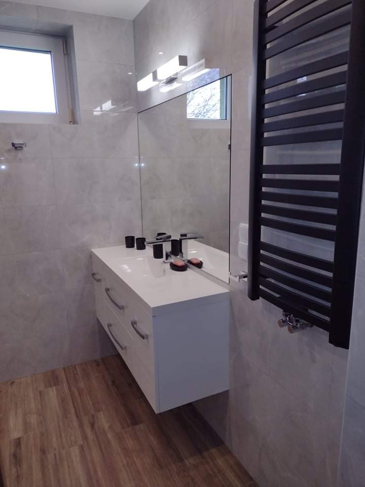 Nowoczesna łazienka z białymi płytkami imitującymi marmur, drewnopodobną podłogą, wiszącą szafką z umywalką i dużym lustrem bez ramy oraz czarnym grzejnikiem drabinkowym.