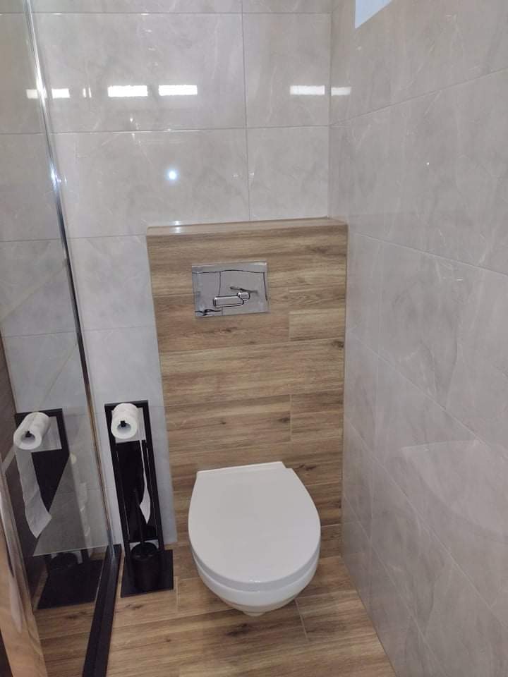 Nowoczesna toaleta w łazience wykończonej płytkami imitującymi marmur i drewno, z widocznym chromowanym przyciskiem spłuczki i czarnym stojakiem na papier toaletowy.