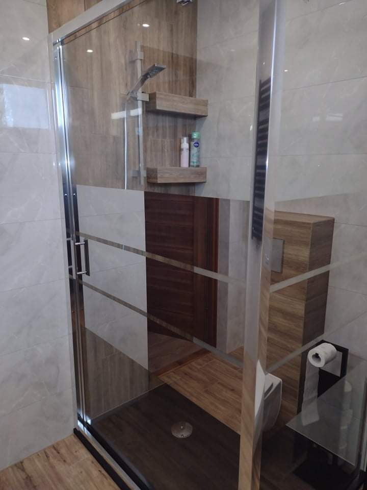 Nowoczesna łazienka z kabiną prysznicową typu walk-in, drewnianymi półkami na kosmetyki, grafitową podłogą i toaletą w zabudowie z drewnopodobnych paneli.
