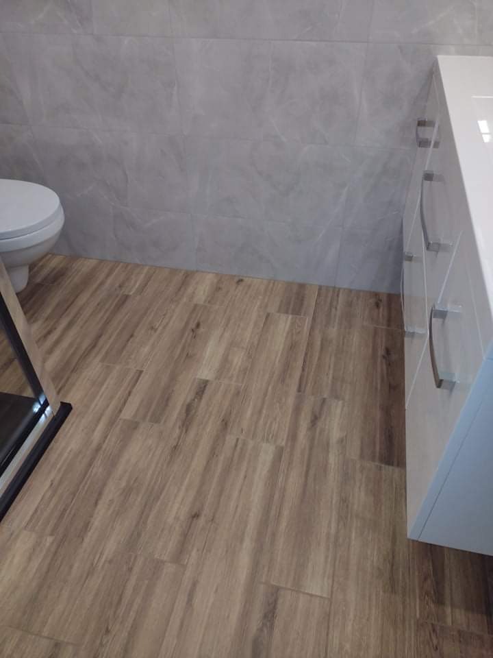 Nowoczesna łazienka z drewnopodobną podłogą, szarymi płytkami na ścianie i białą szafką z chromowanymi uchwytami, widoczny fragment toalety i kabiny prysznicowej.