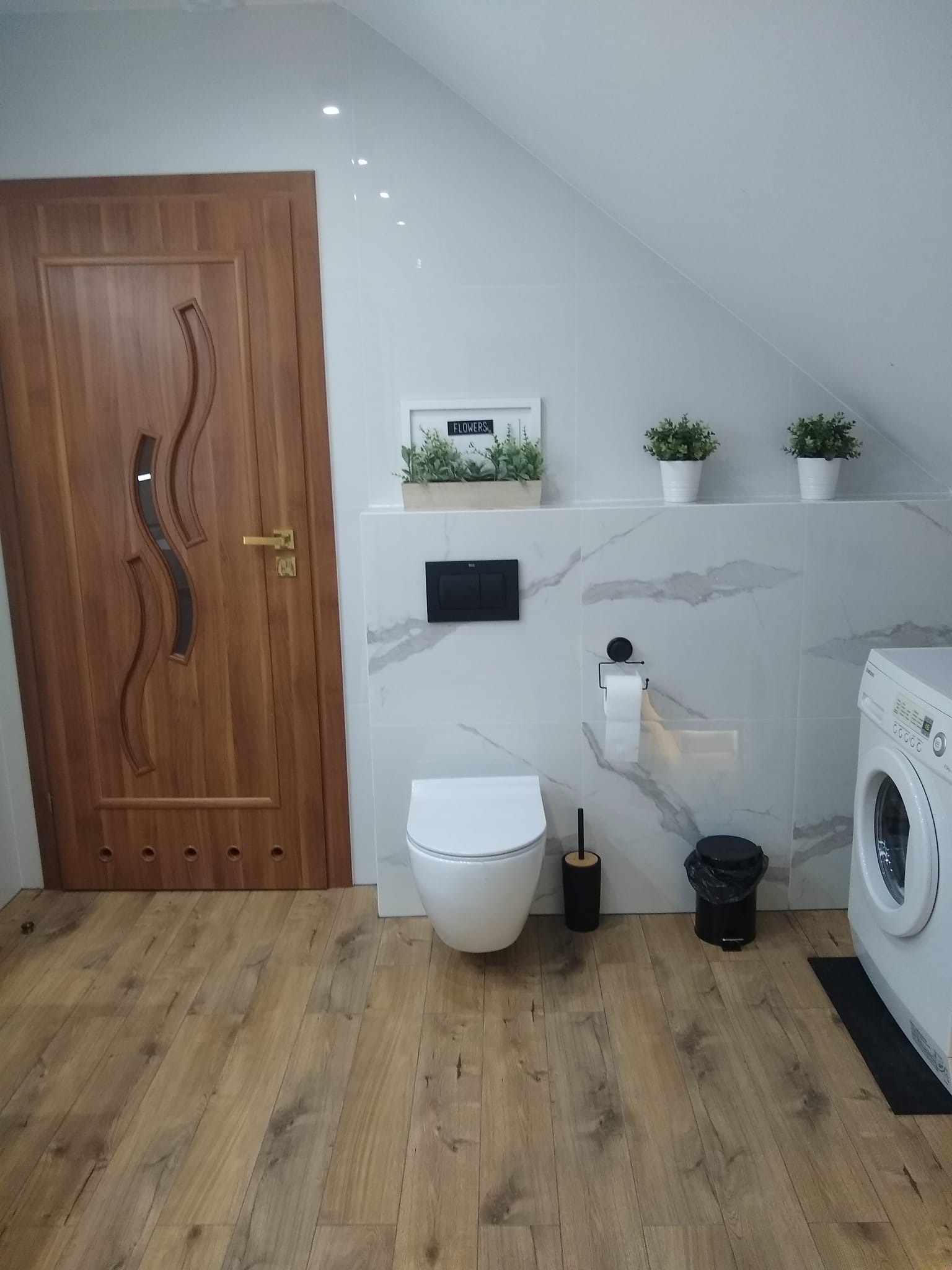 Nowoczesna aranżacja łazienki na poddaszu w Tarnowie, z drewnianą podłogą, marmurowymi płytkami ściennymi, wiszącą toaletą i pralką.