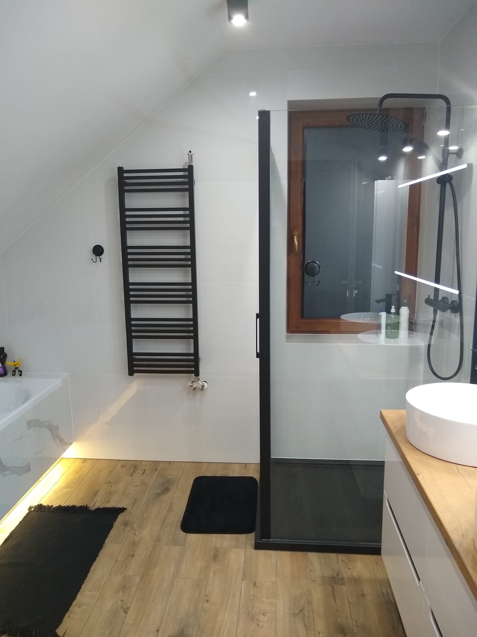 Nowoczesna łazienka na poddaszu z drewnianą podłogą, białymi płytkami, czarnym grzejnikiem drabinkowym, kabiną prysznicową typu walk-in z czarną ramą, oknem w drewnianej ramie i oświetleniem LED...