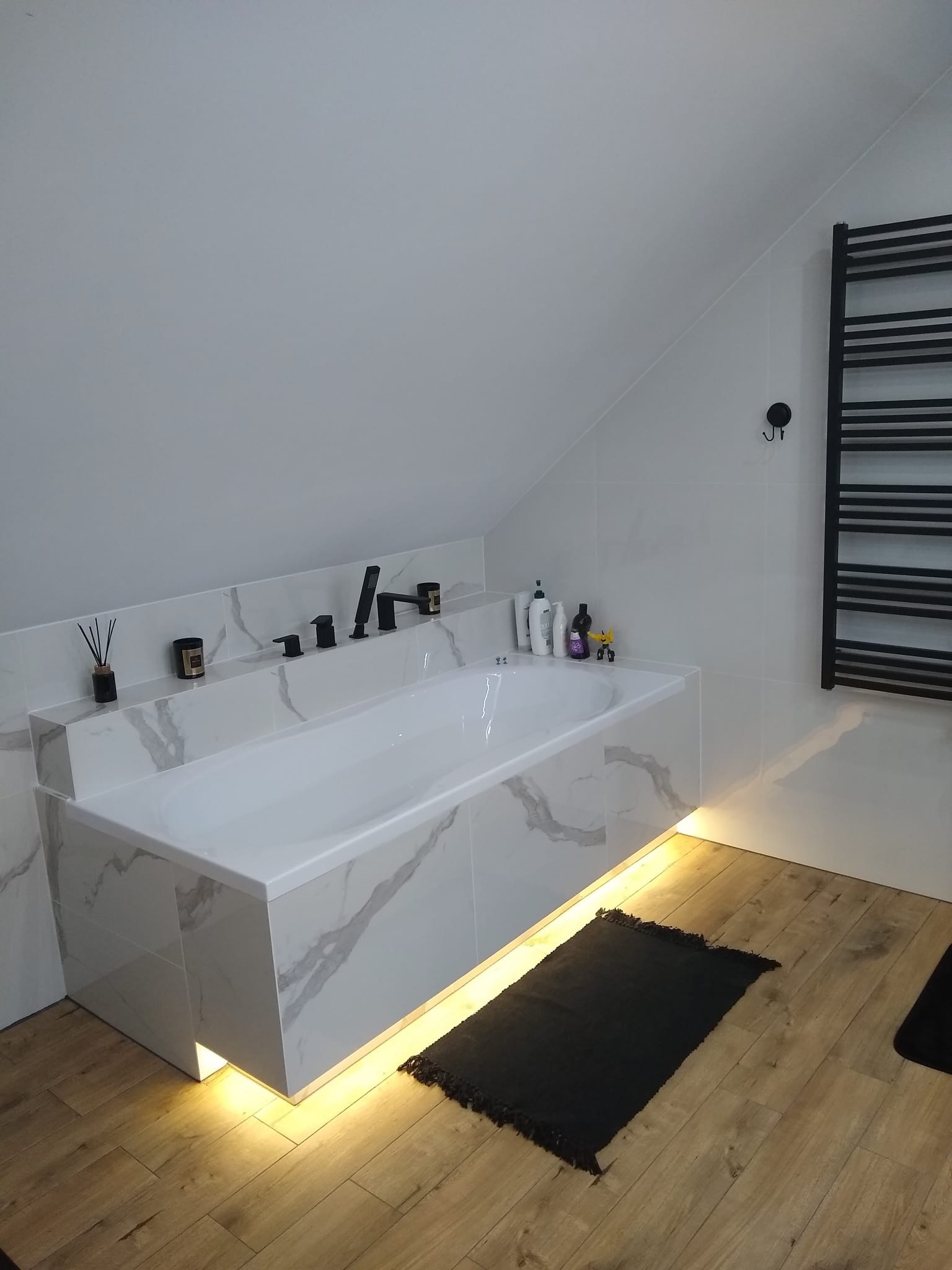 Biała wanna obudowana płytkami imitującymi marmur, z czarną armaturą i podświetleniem LED, w łazience na poddaszu z drewnianą podłogą i czarnym grzejnikiem drabinkowym.