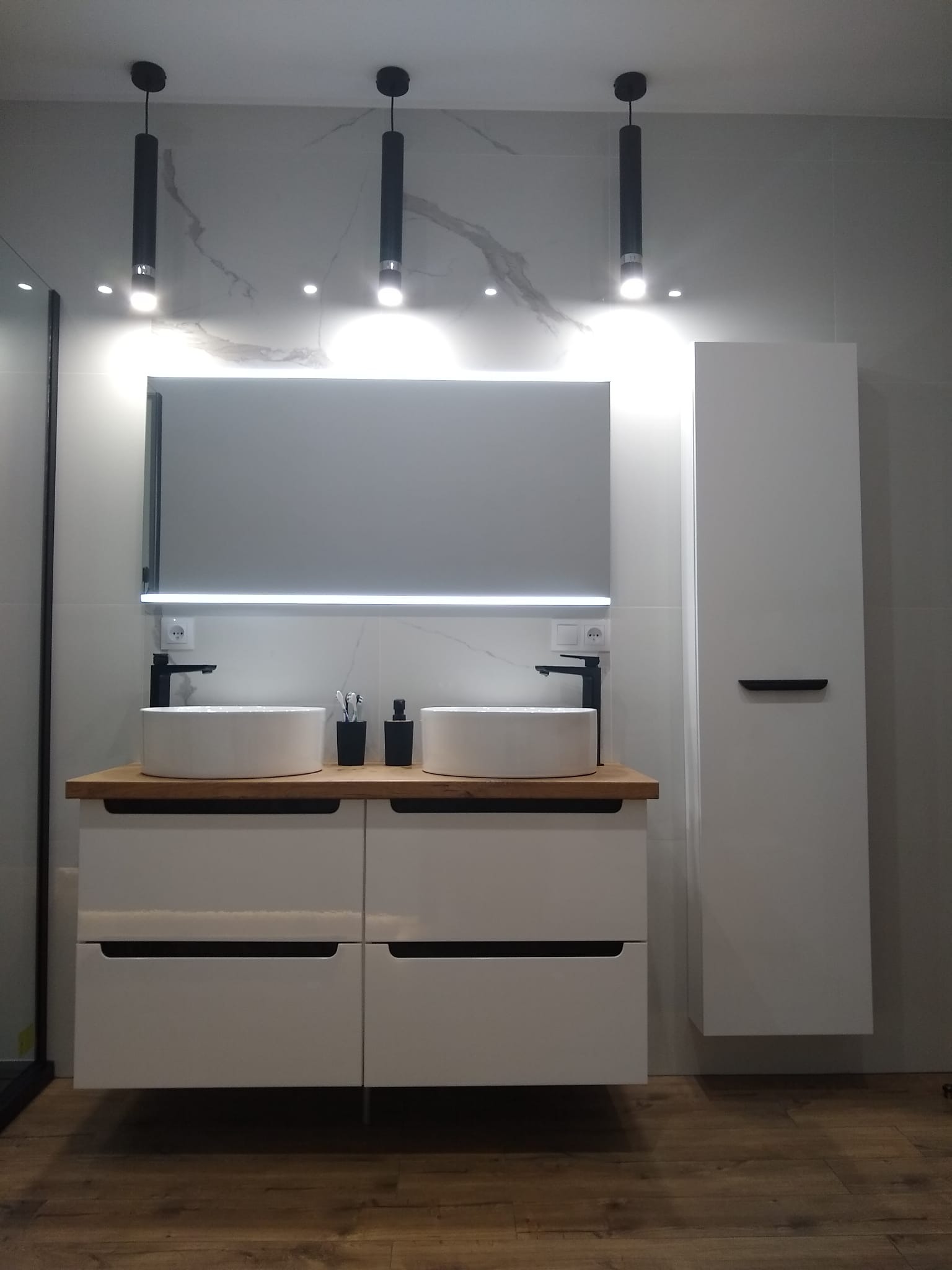 Nowoczesna łazienka z dwiema umywalkami nablatowymi, czarną armaturą, podświetlanym lustrem i wiszącą szafką, wykończona płytkami imitującymi marmur.