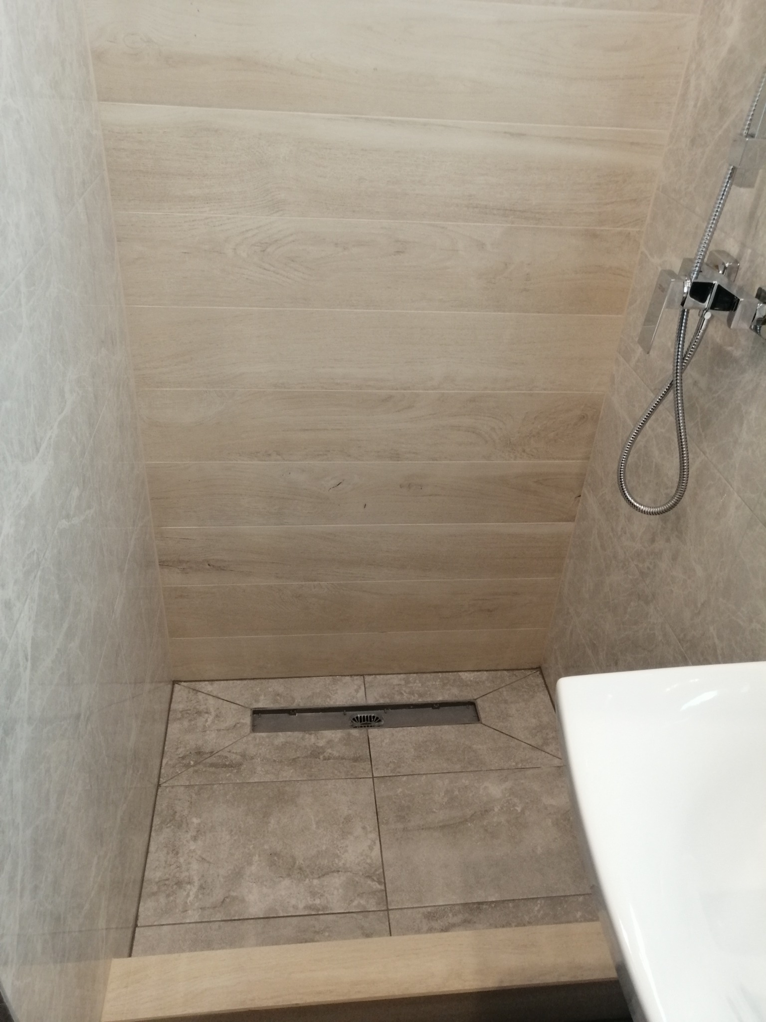 Wykończony brodzik kabiny prysznicowej z odpływem liniowym, wyłożony płytkami w odcieniach szarości i beżu, z widoczną baterią prysznicową i fragmentem umywalki.