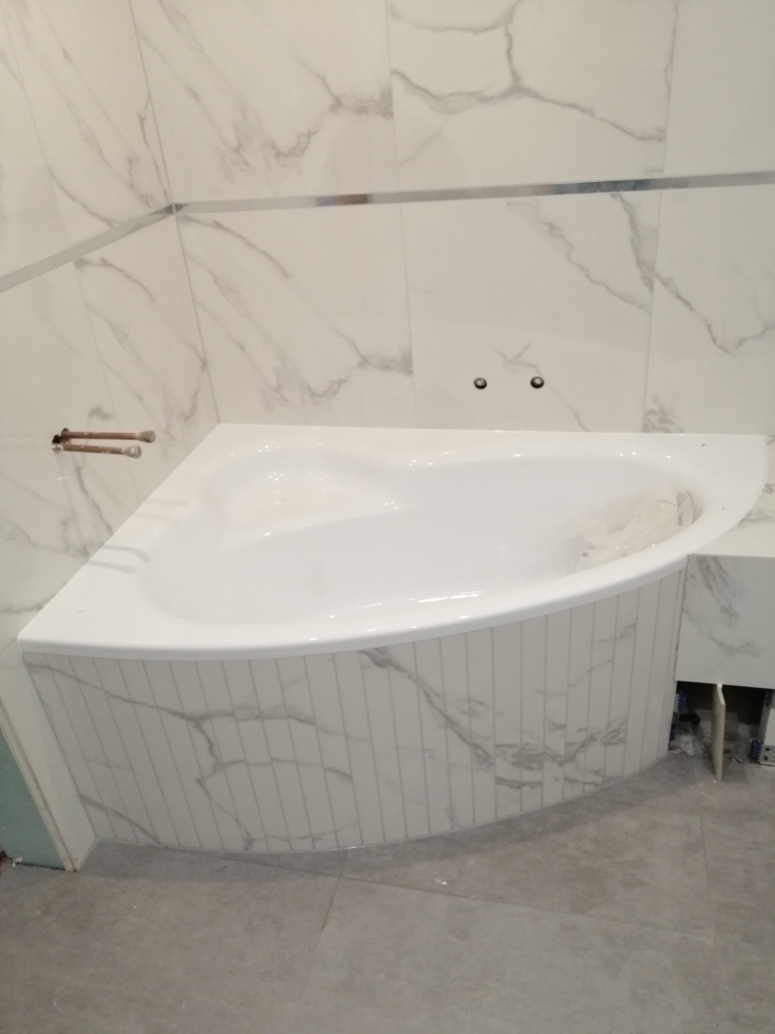Narożna wanna obudowana płytkami imitującymi marmur, z widocznymi wystającymi rurami instalacji wodnej i fragmentem niezakończonej obudowy szafki pod umywalkę, na podłodze szare płytki.