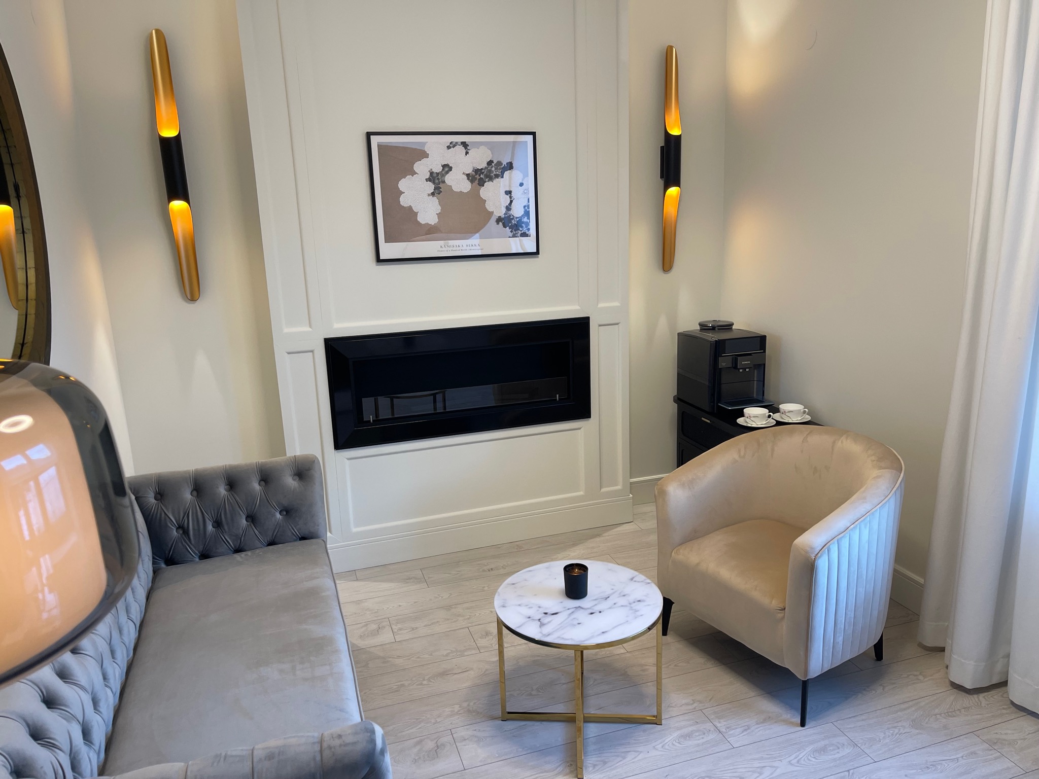 Elegancki gabinet z szarą sofą Chesterfield, beżowym fotelem, marmurowym stolikiem kawowym i nowoczesnym kominkiem elektrycznym.