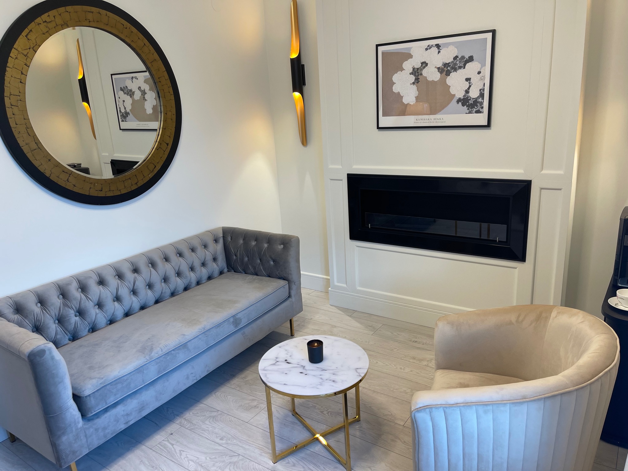 Wnętrze gabinetu z szarym, pikowanym siedziskiem, okrągłym lustrem w złotej ramie, marmurowym stolikiem kawowym z czarną świecą i beżowym fotelem na tle białych ścian z czarnym kominkiem...