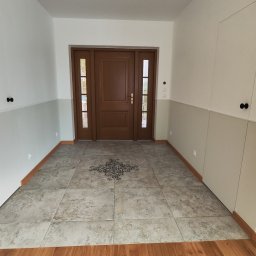 Andrzej Sarkowicz Remonty i wykończenia - Przedpokój po remoncie: szare płytki podłogowe z dekoracyjnym wzorem centralnym, brązowe drewniane drzwi wejściowe, ściany w dwóch odcieniach szarości z białymi szafkami w zabudowie.