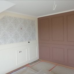 Andrzej Sarkowicz Remonty i wykończenia - Pokój po częściowym remoncie: ściana z beżową tapetą w ornamenty i białą boazerią, sąsiednia ściana w odcieniach brązu z dekoracyjnymi panelami, podłoga zabezpieczona folią malarską.