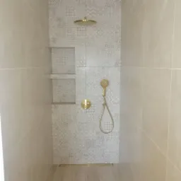 Wykończona kabina prysznicowa z szarymi płytkami o zróżnicowanym wzorze, złotą armaturą i wnękami na kosmetyki.