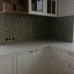 Fragment kuchni z oliwkowymi płytkami dekoracyjnymi na ścianie nad białym blatem i białymi szafkami z ozdobnymi uchwytami; widoczne puszki elektryczne przed montażem gniazdek.