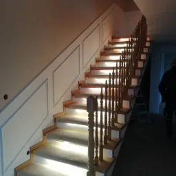 Drewniane schody z białymi podstopnicami i balustradą, zabezpieczone folią malarską podczas prac wykończeniowych, ściana z dekoracyjnymi panelami.