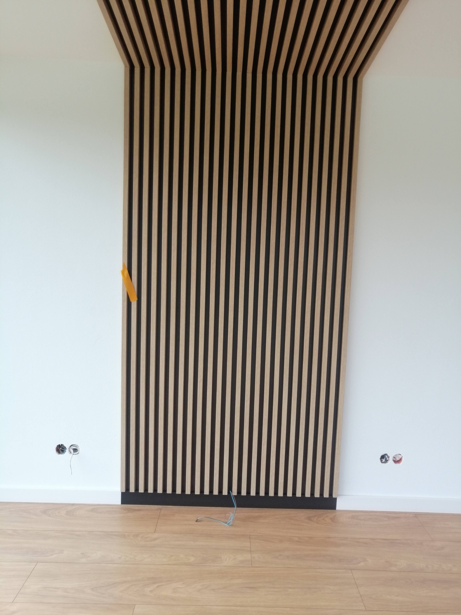 Drewniane lamele ścienne w kolorze jasnego drewna i czerni, zamontowane na białej ścianie, z widocznymi gniazdkami elektrycznymi i fragmentem podłogi z paneli.