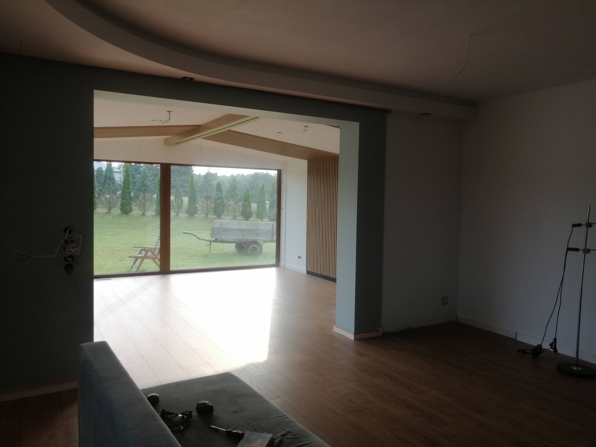 Wnętrze domu w trakcie prac wykończeniowych: świeżo pomalowane ściany w odcieniach szarości, drewniana podłoga, widoczne instalacje elektryczne i duże okno z widokiem na ogród z przyczepką.