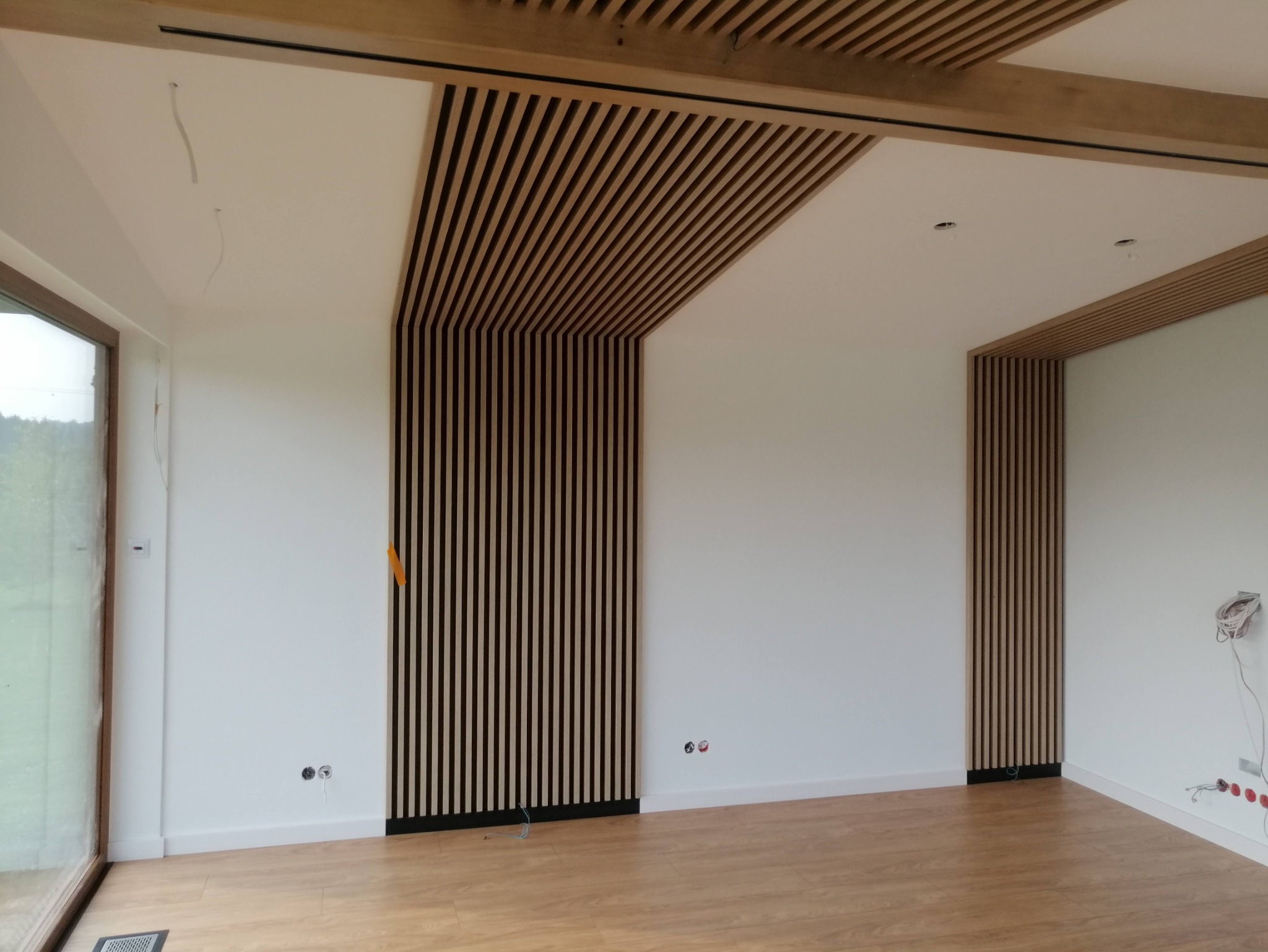 Nowoczesne wykończenie wnętrza z drewnianymi lamelami na suficie i ścianach, widoczne niedokończone instalacje elektryczne i fragment okna.