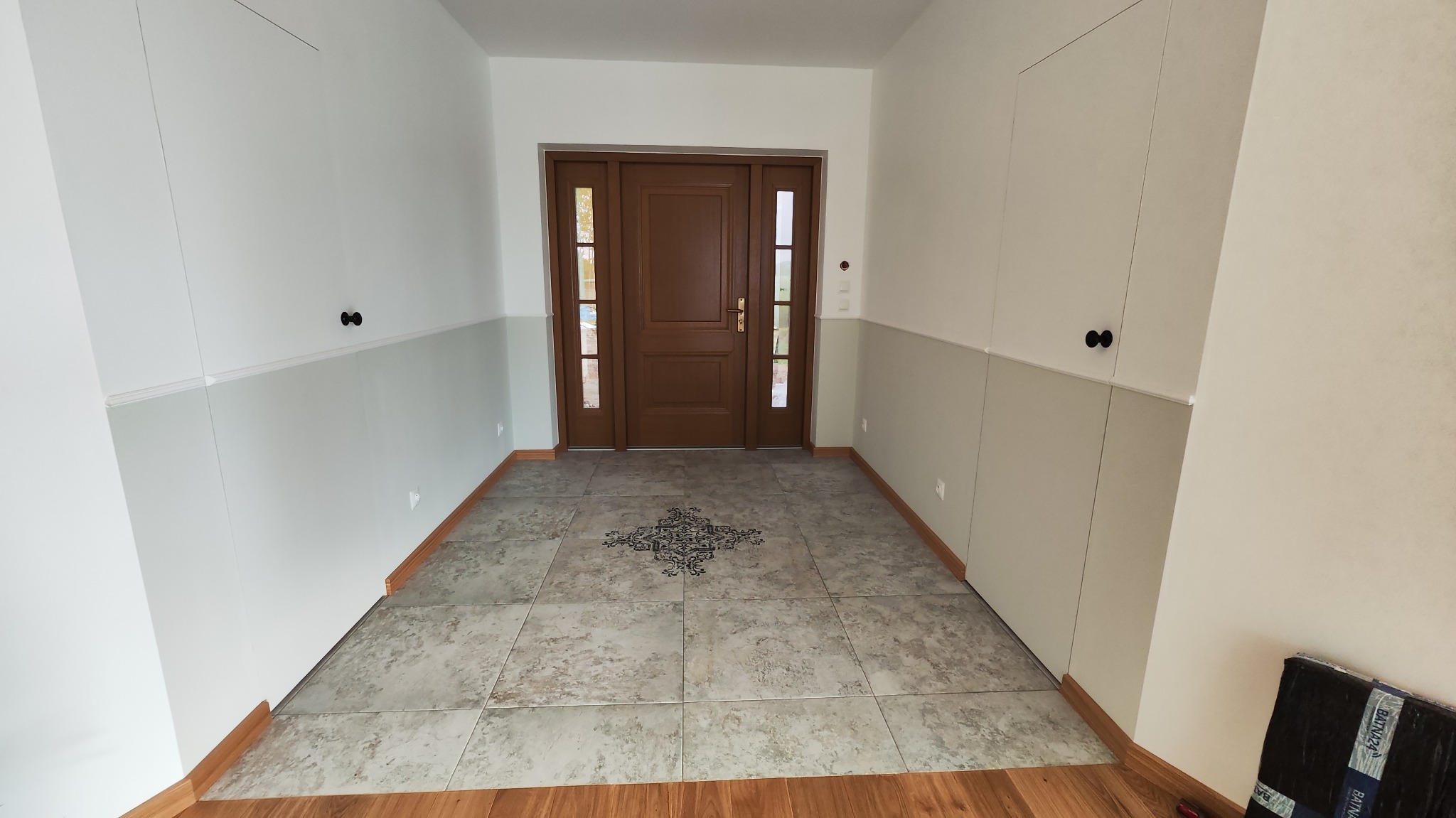 Przedpokój po remoncie: szare płytki podłogowe z dekoracyjnym wzorem centralnym, brązowe drewniane drzwi wejściowe, ściany w dwóch odcieniach szarości z białymi szafkami w zabudowie.