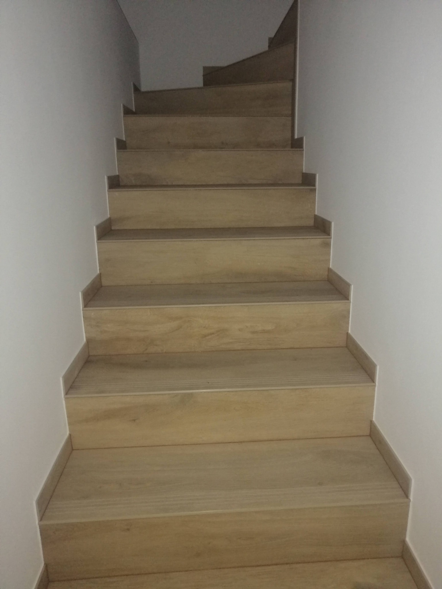 Klatka schodowa wyłożona płytkami imitującymi jasne drewno, z widocznymi listwami przypodłogowymi i białymi ścianami, ujęcie z dołu.