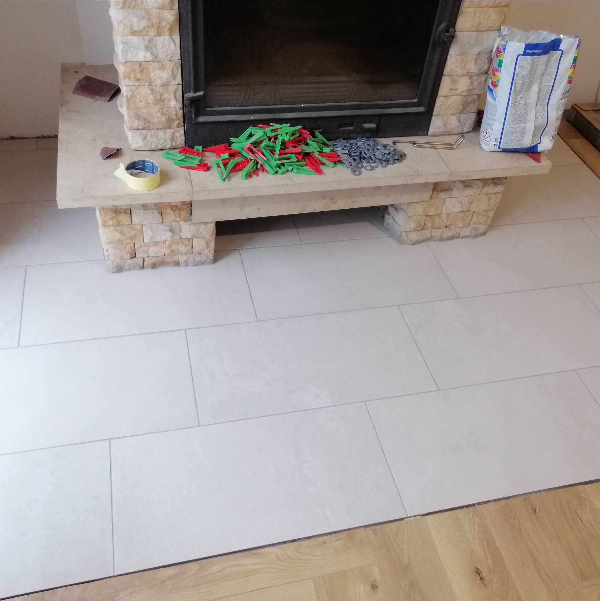 Jasne, prostokątne płytki podłogowe ułożone przy kamiennym obramowaniu kominka, widoczne narzędzia do układania płytek: kliny, poziomice i worek z klejem.
