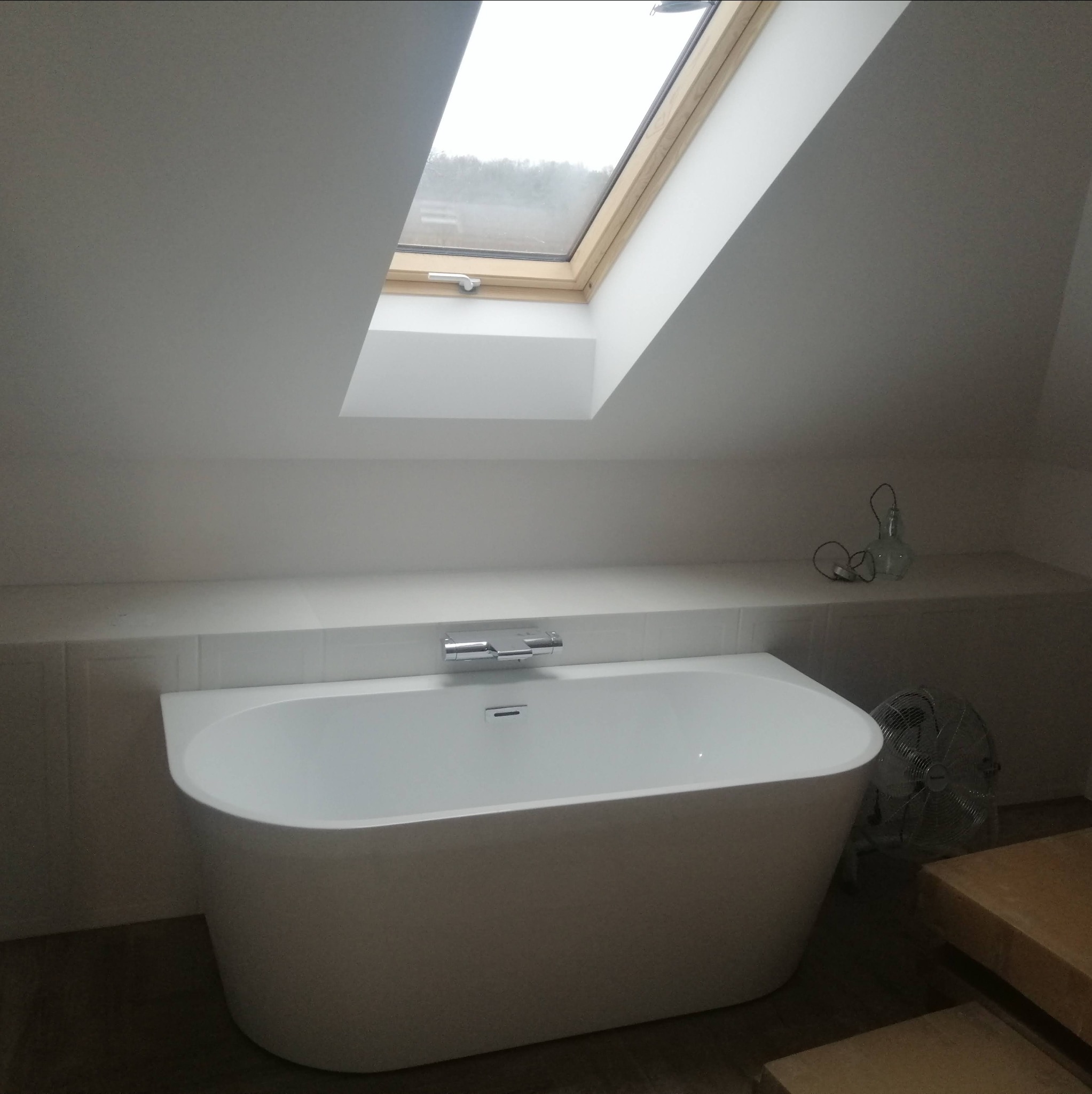 Wolnostojąca wanna w jasnej łazience na poddaszu z oknem dachowym i półką z dekoracjami, widoczny fragment podłogi i wentylator.