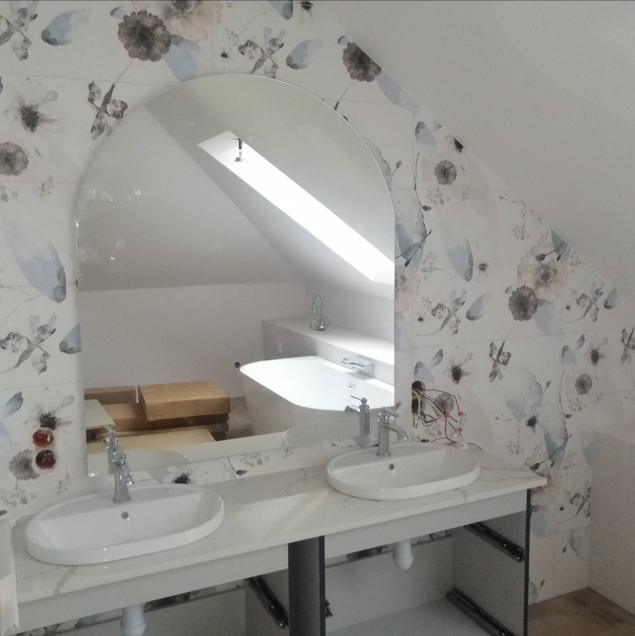 Umywalka nablatowa z baterią, lustro łukowe na tle ściany w kwiatowe płytki, widoczna wanna i okno dachowe w odbiciu, instalacja elektryczna w trakcie podłączania.