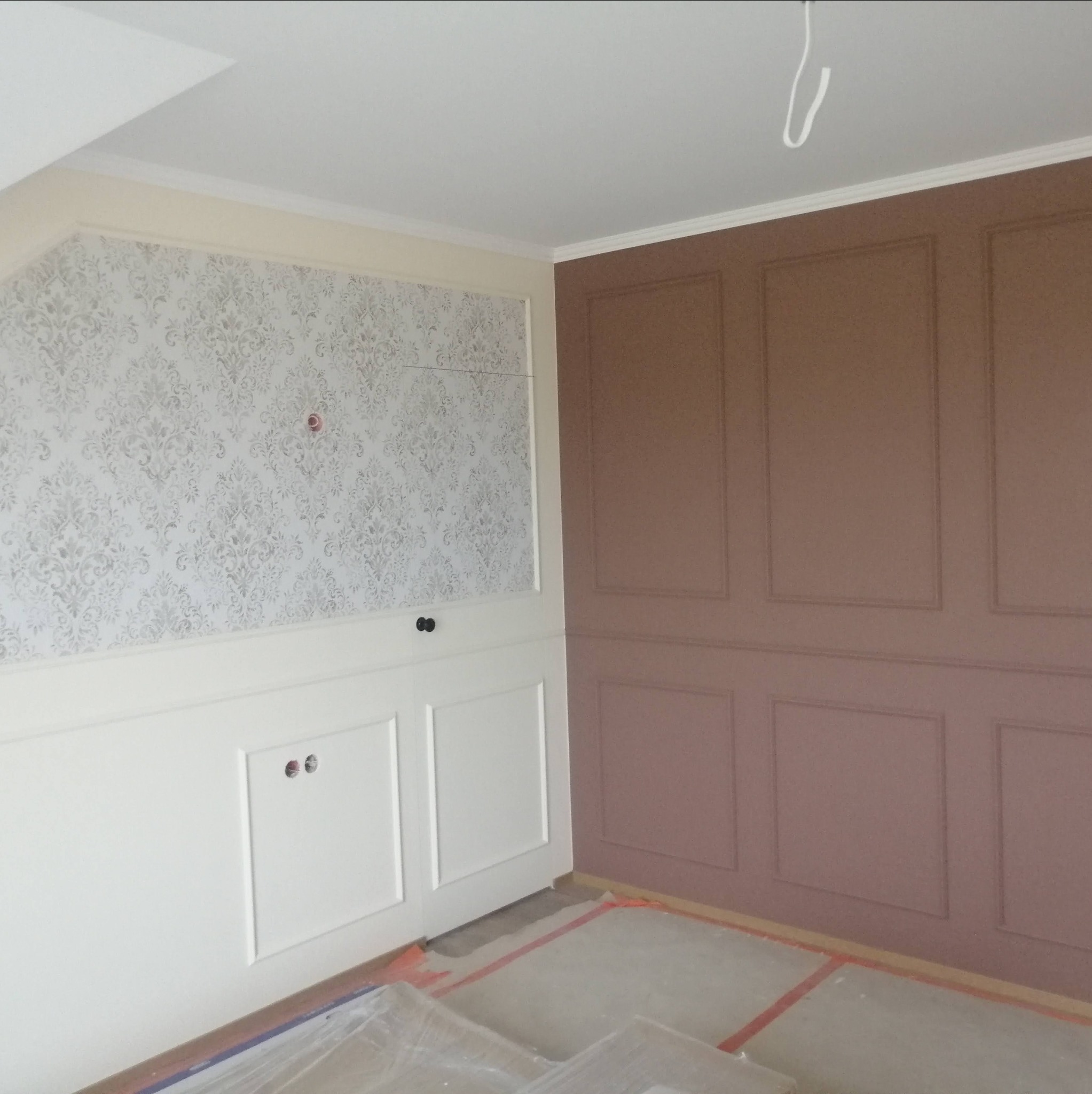 Pokój po częściowym remoncie: ściana z beżową tapetą w ornamenty i białą boazerią, sąsiednia ściana w odcieniach brązu z dekoracyjnymi panelami, podłoga zabezpieczona folią malarską.