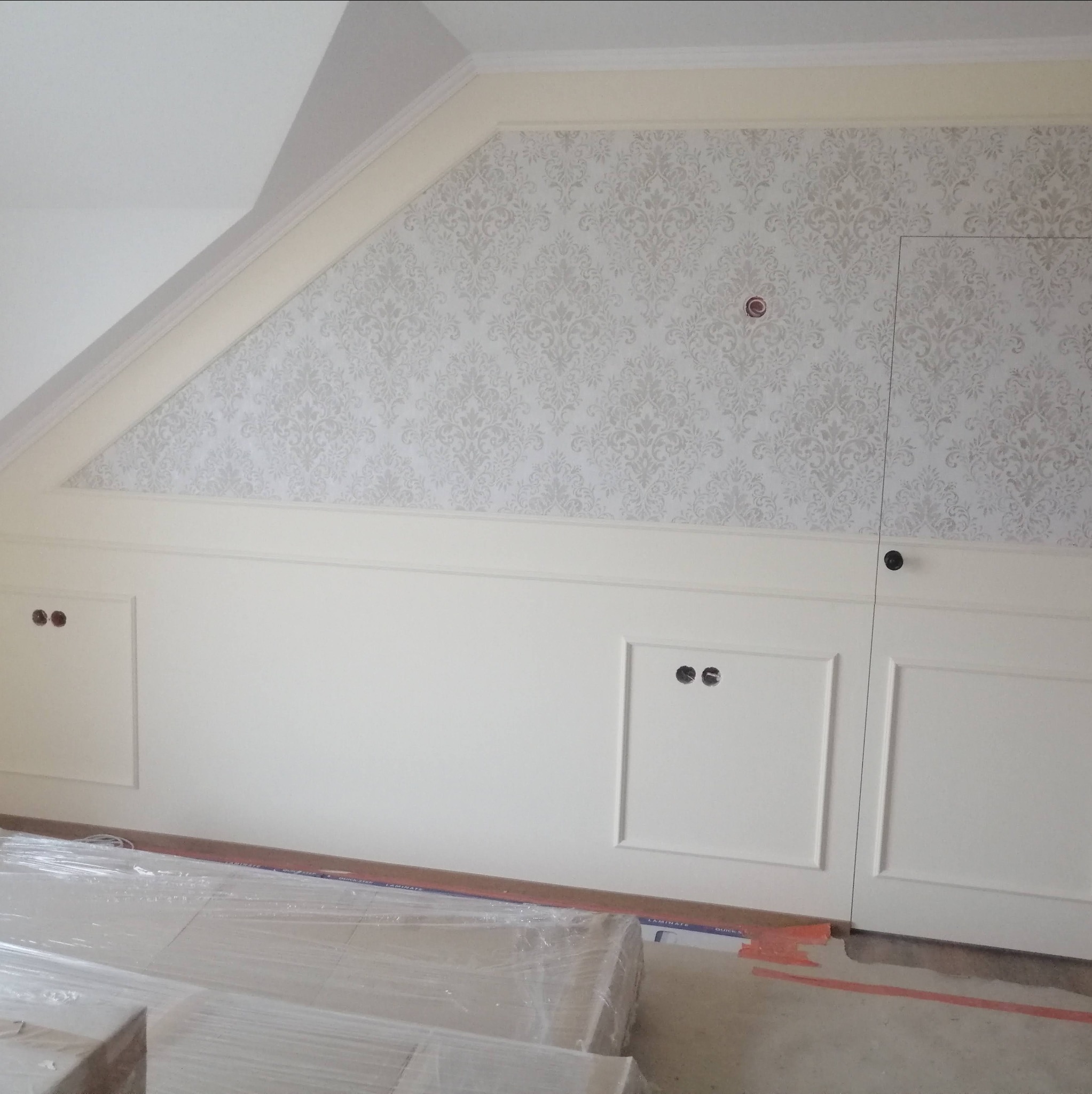 Ściana pod skosem z tapetą w ornamenty, biała boazeria z ukrytymi drzwiczkami i przygotowane do montażu panele podłogowe w folii.