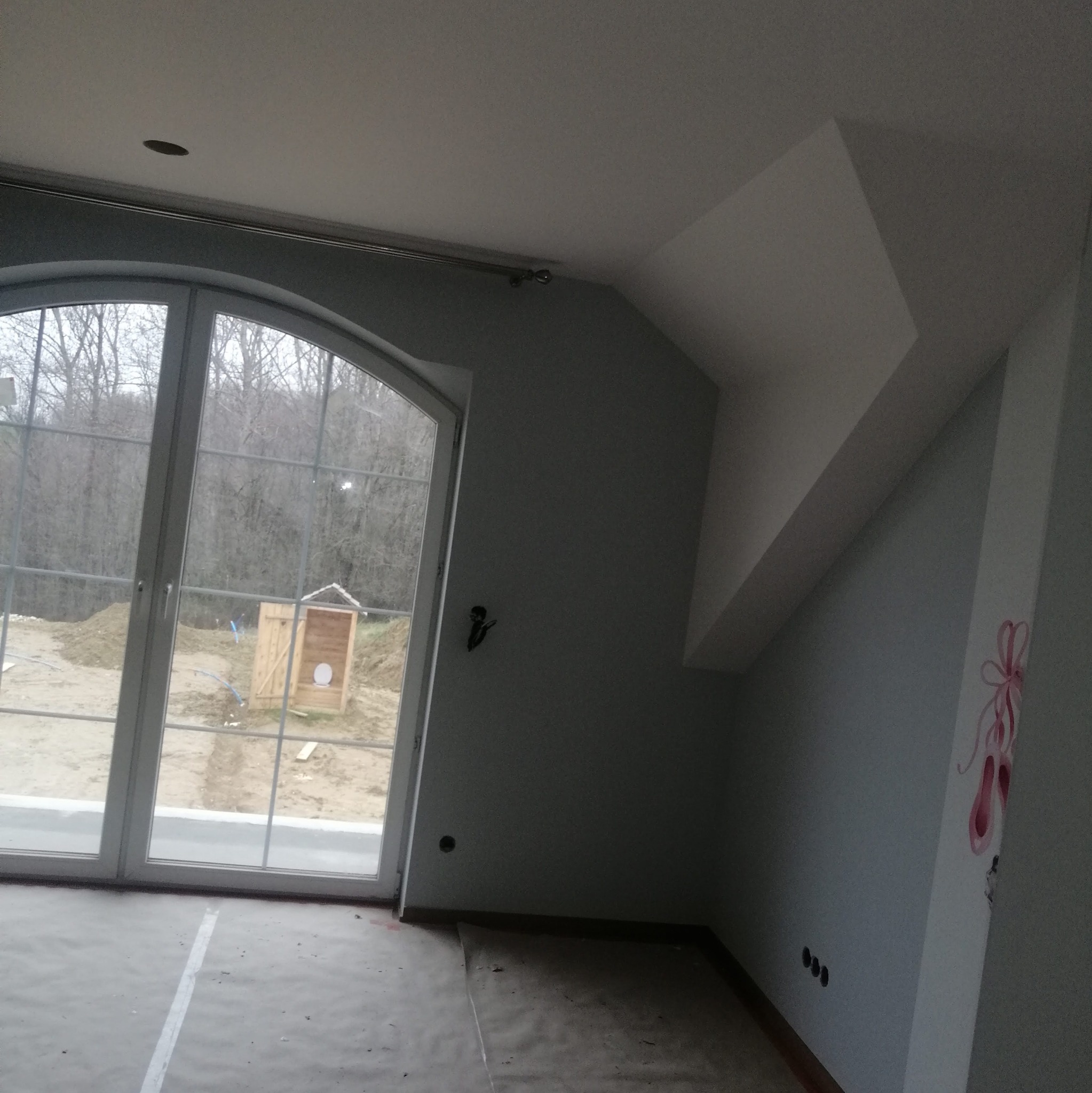 Wnętrze pokoju w trakcie prac wykończeniowych: ściany pomalowane na szaro, widoczne malunki baletek na ścianie, okno z widokiem na zewnątrz, podłoga zabezpieczona folią malarską, surowy stan.