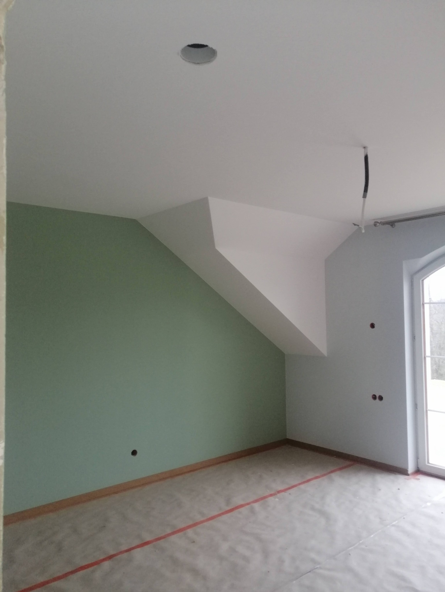 Pokój na poddaszu po malowaniu ścian na zielono i szaro, z widocznym otworem na oświetlenie w suficie i zabezpieczeniem podłogi folią malarską.