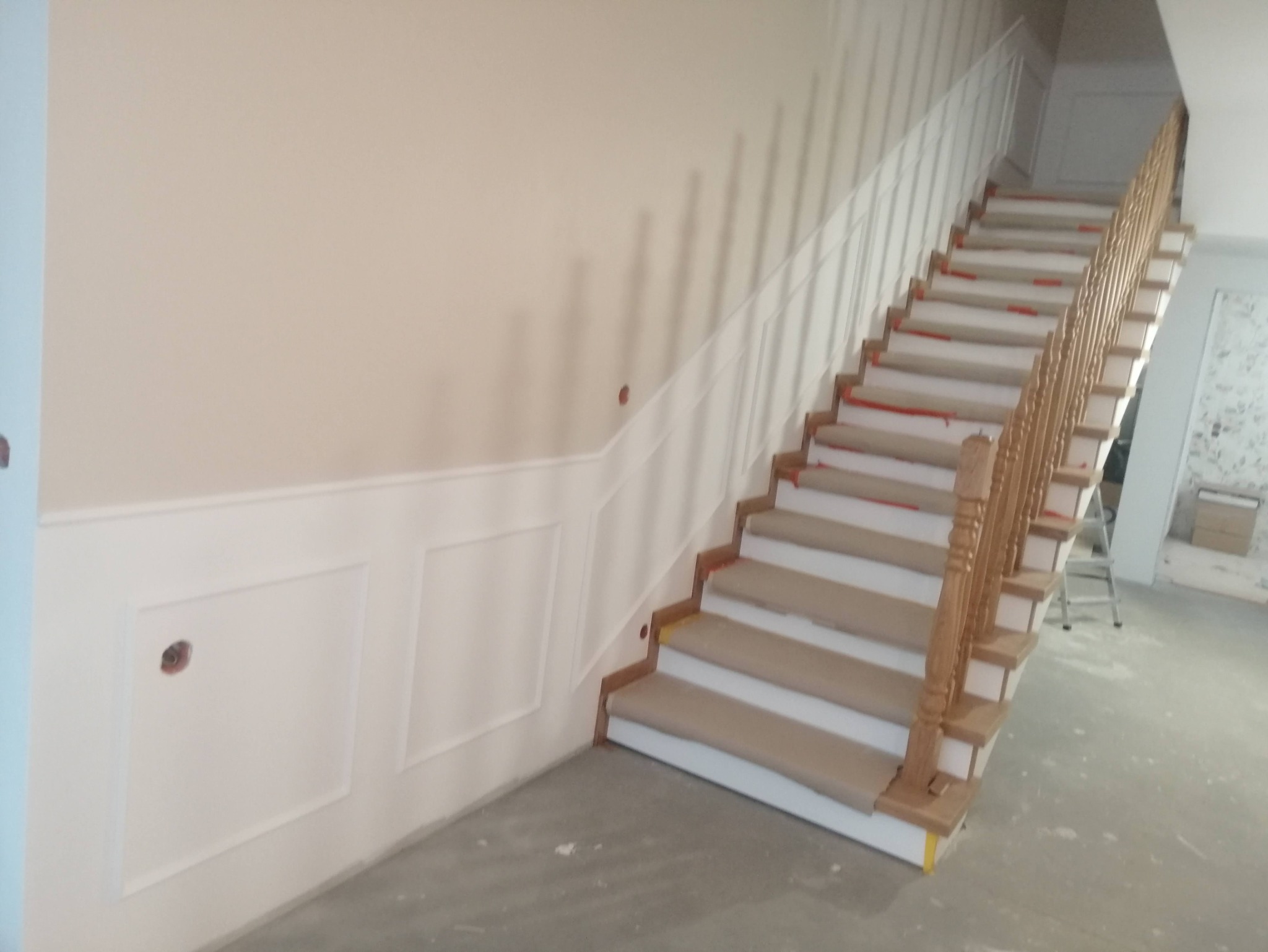 Klatka schodowa w trakcie prac wykończeniowych, zabezpieczone stopnie, listwy dekoracyjne na ścianie, widoczne ślady po gniazdkach elektrycznych.