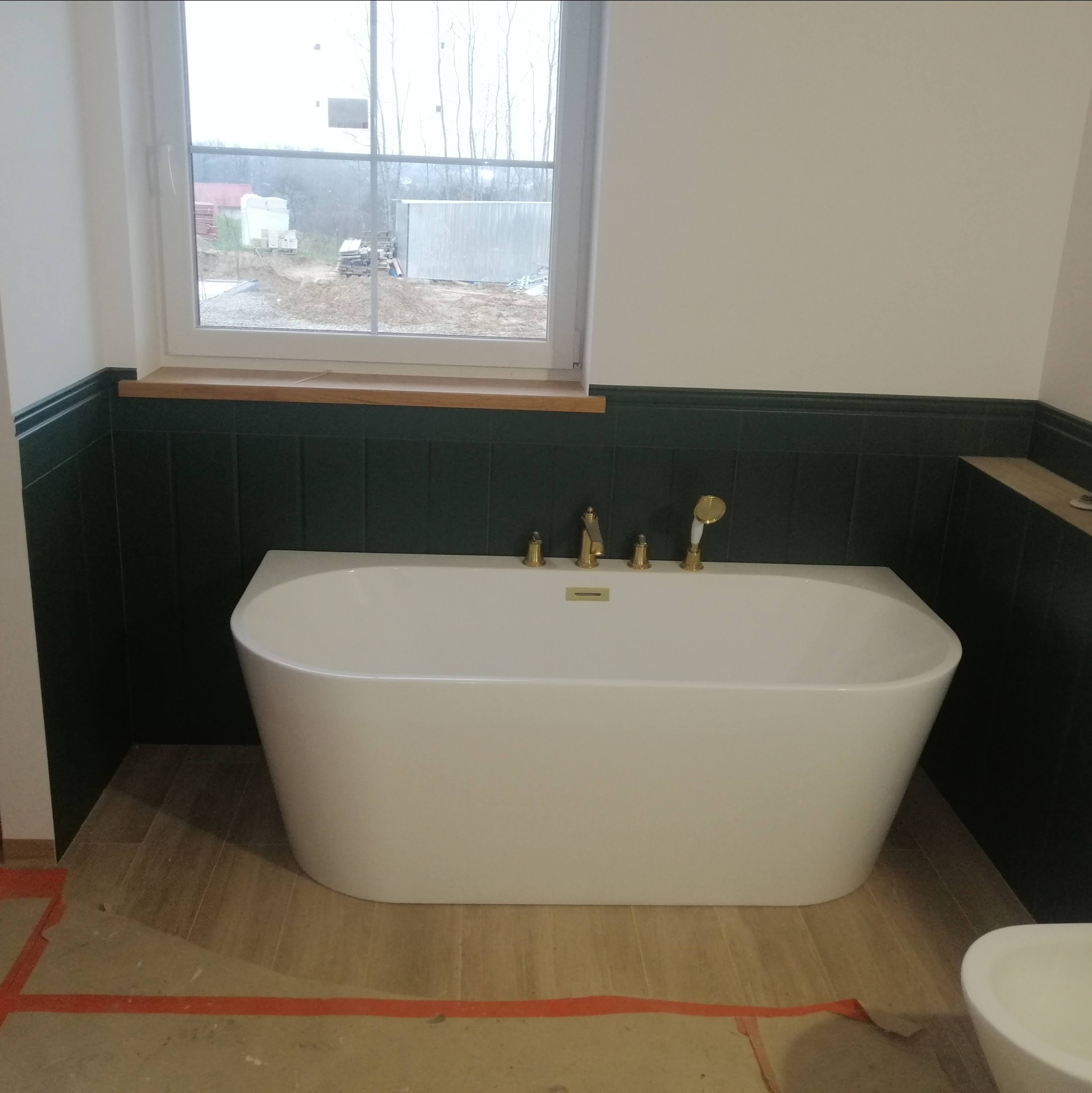 Wolnostojąca wanna z baterią w stylu retro na tle ciemnozielonych płytek i drewnianego parapetu okiennego; widoczne zabezpieczenia podłogi.
