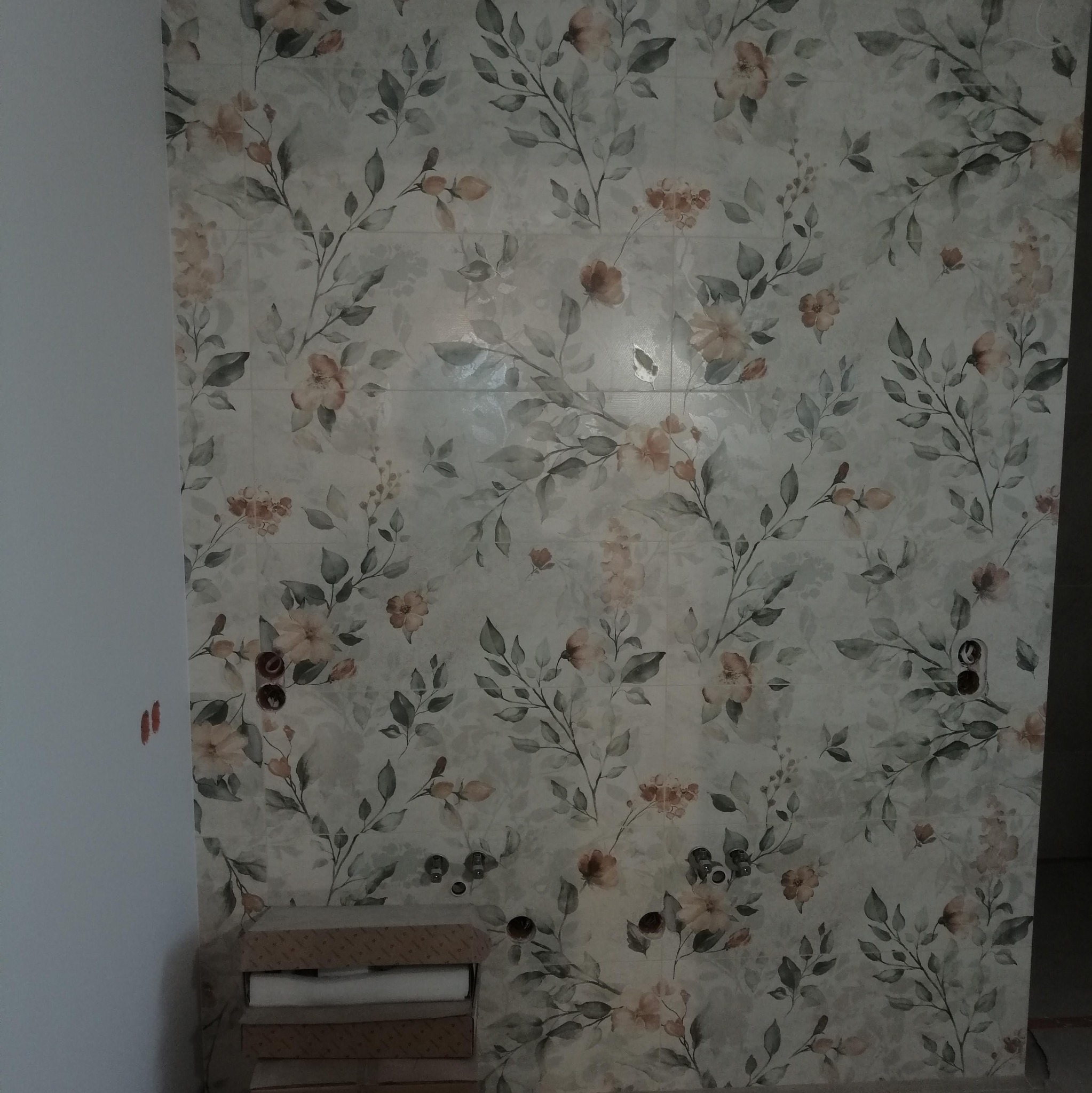 Ściana wyłożona płytkami z motywem kwiatowym, widoczne puszki elektryczne i rury hydrauliczne, na podłodze stos kartonów z płytkami.