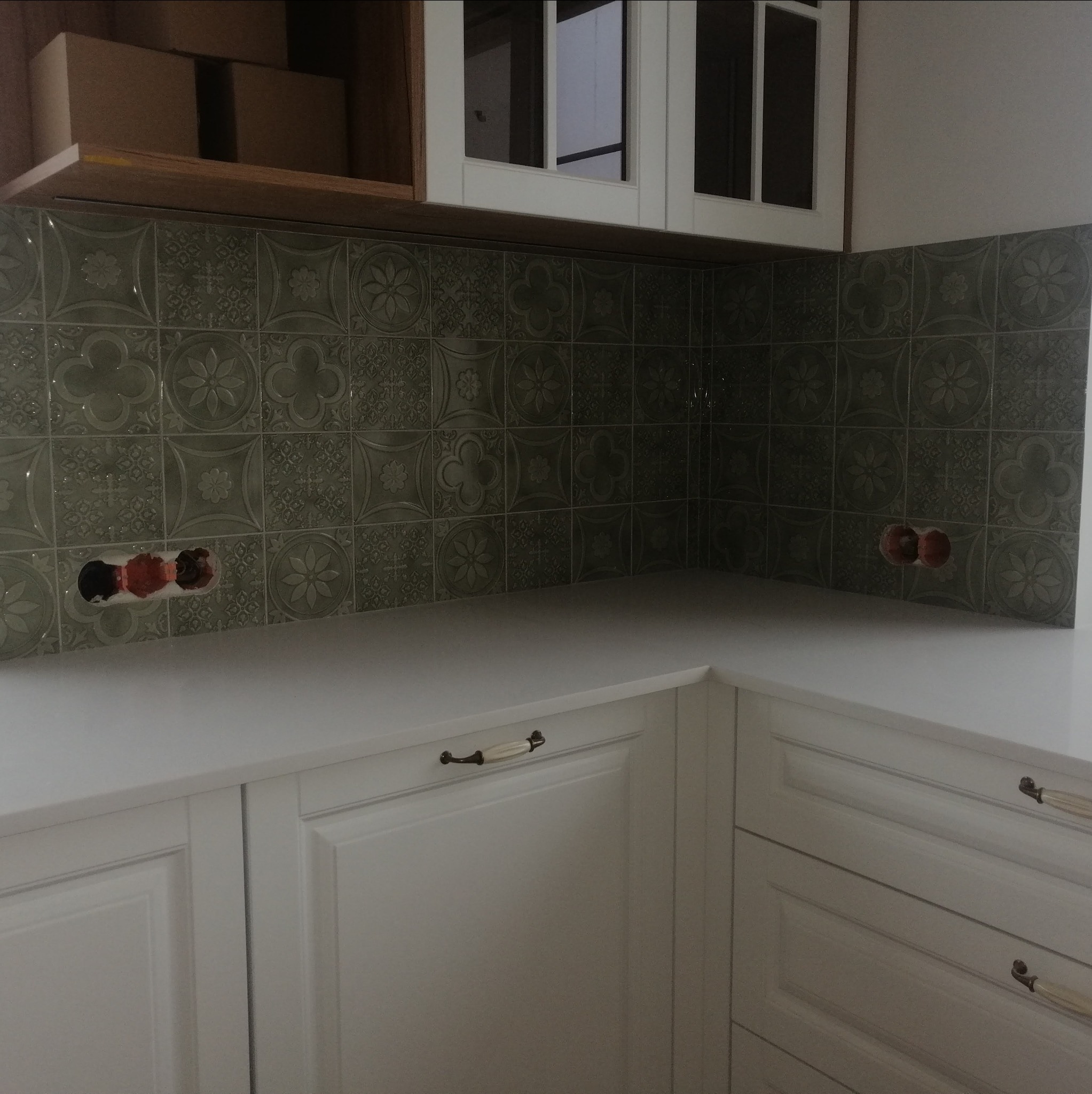 Fragment kuchni z oliwkowymi płytkami dekoracyjnymi na ścianie nad białym blatem i białymi szafkami z ozdobnymi uchwytami; widoczne puszki elektryczne przed montażem gniazdek.