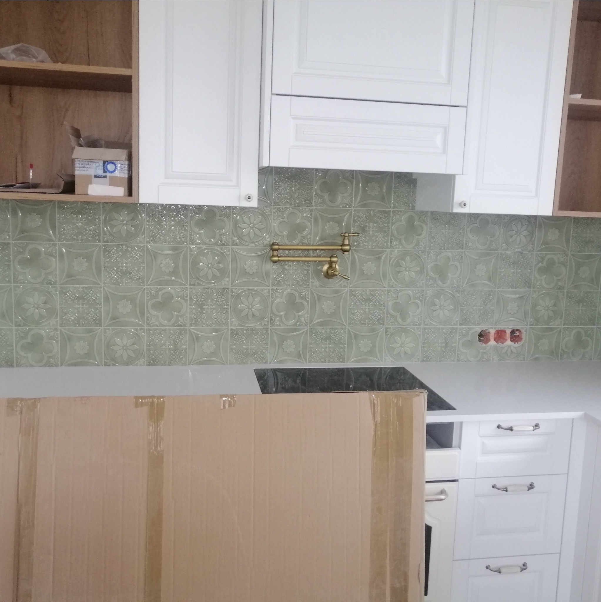 Montaż kuchni: białe szafki wiszące i dolne z dekoracyjnymi uchwytami, szare, wzorzyste płytki na ścianie, bateria kuchenna w kolorze złota, widoczne puszki elektryczne. Część kuchni zasłonięta...