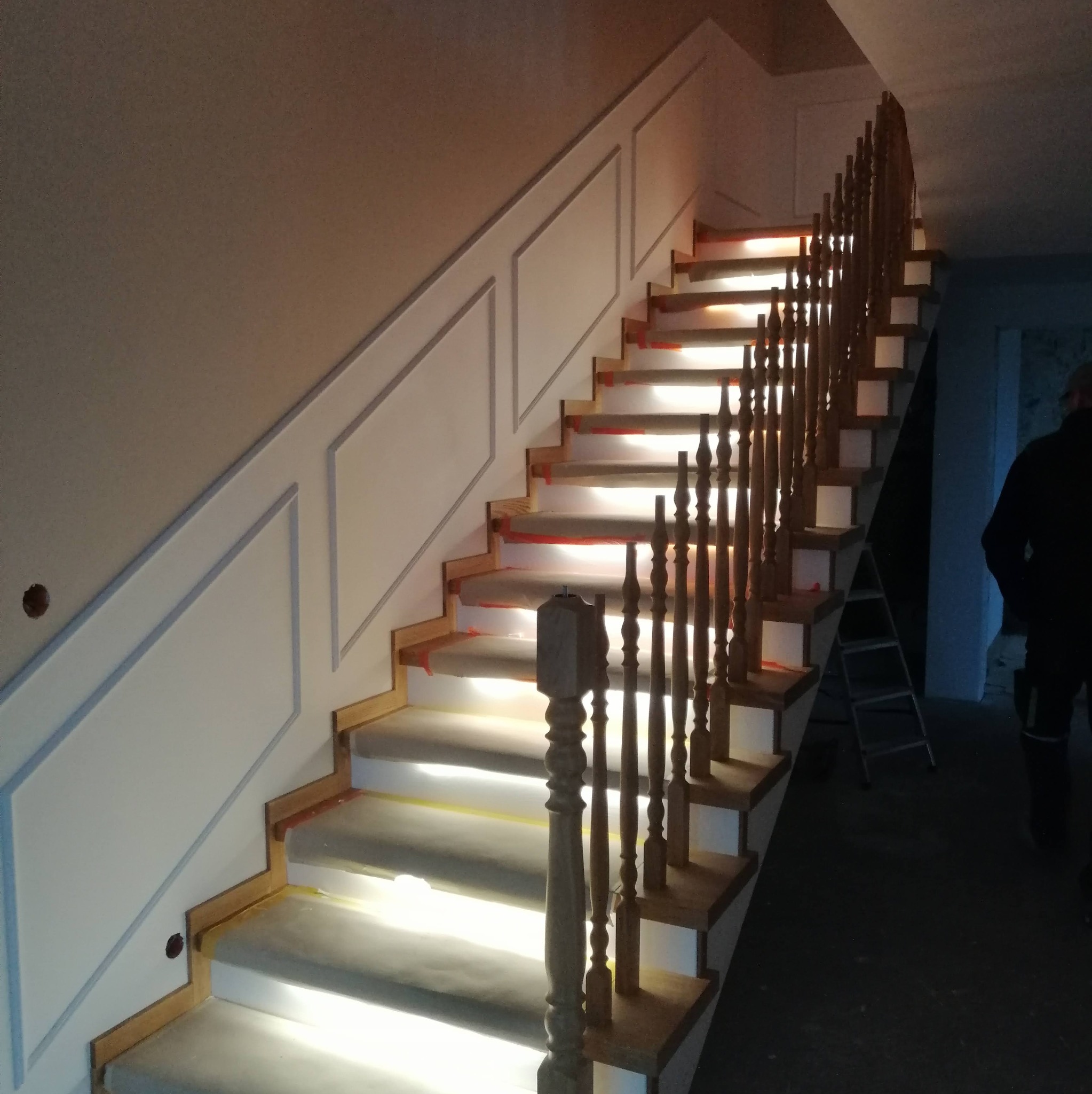 Drewniane schody z białymi podstopnicami i balustradą, zabezpieczone folią malarską podczas prac wykończeniowych, ściana z dekoracyjnymi panelami.