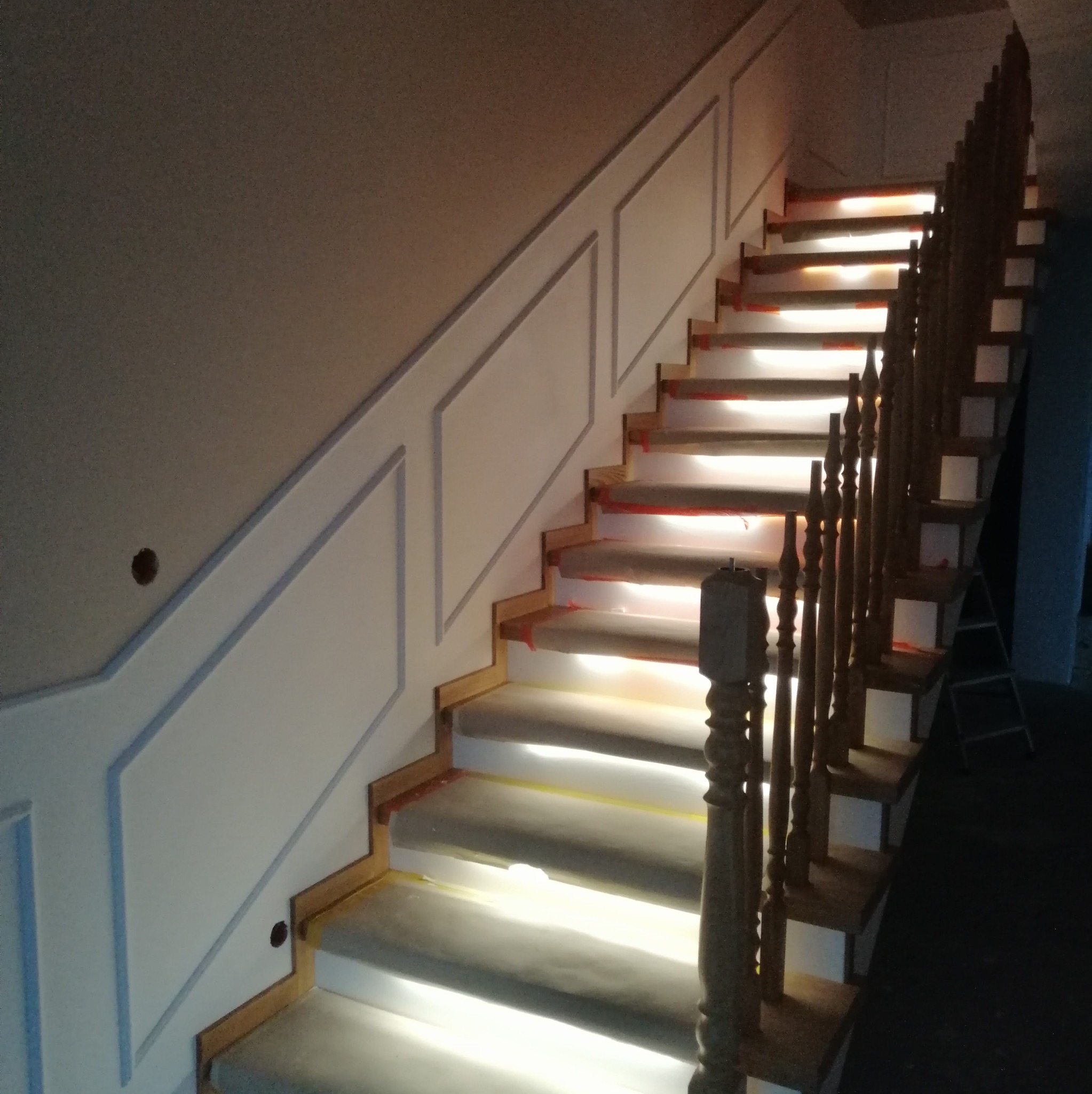 Schody drewniane z białymi panelami ściennymi podczas montażu oświetlenia LED pod stopniami, zabezpieczone folią malarską.