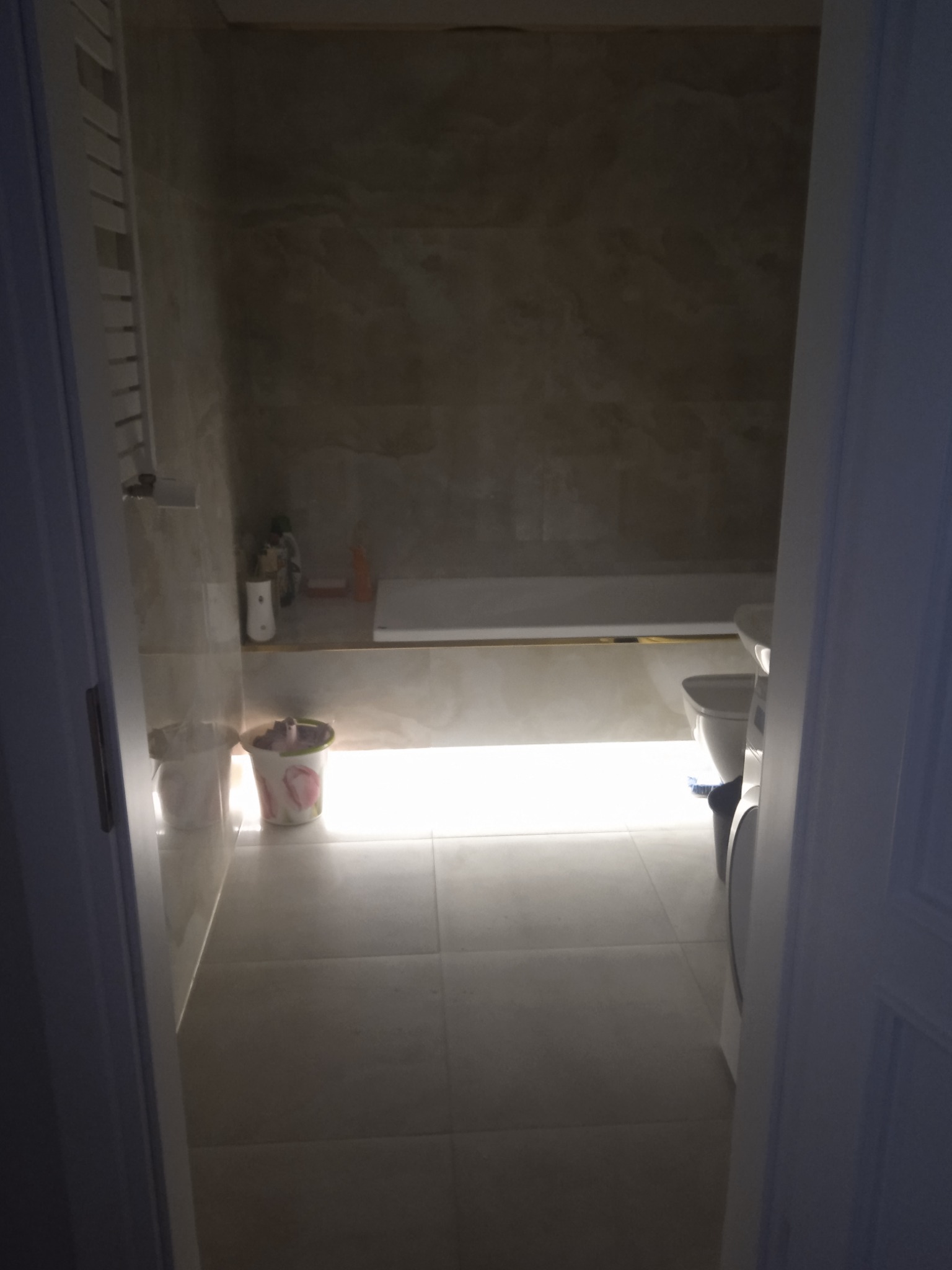 Widok zza uchylonych drzwi na łazienkę z beżowymi płytkami, białą wanną z złotym wykończeniem, i toaletą. Na podłodze stoi kosz na śmieci z motywem kwiatowym.