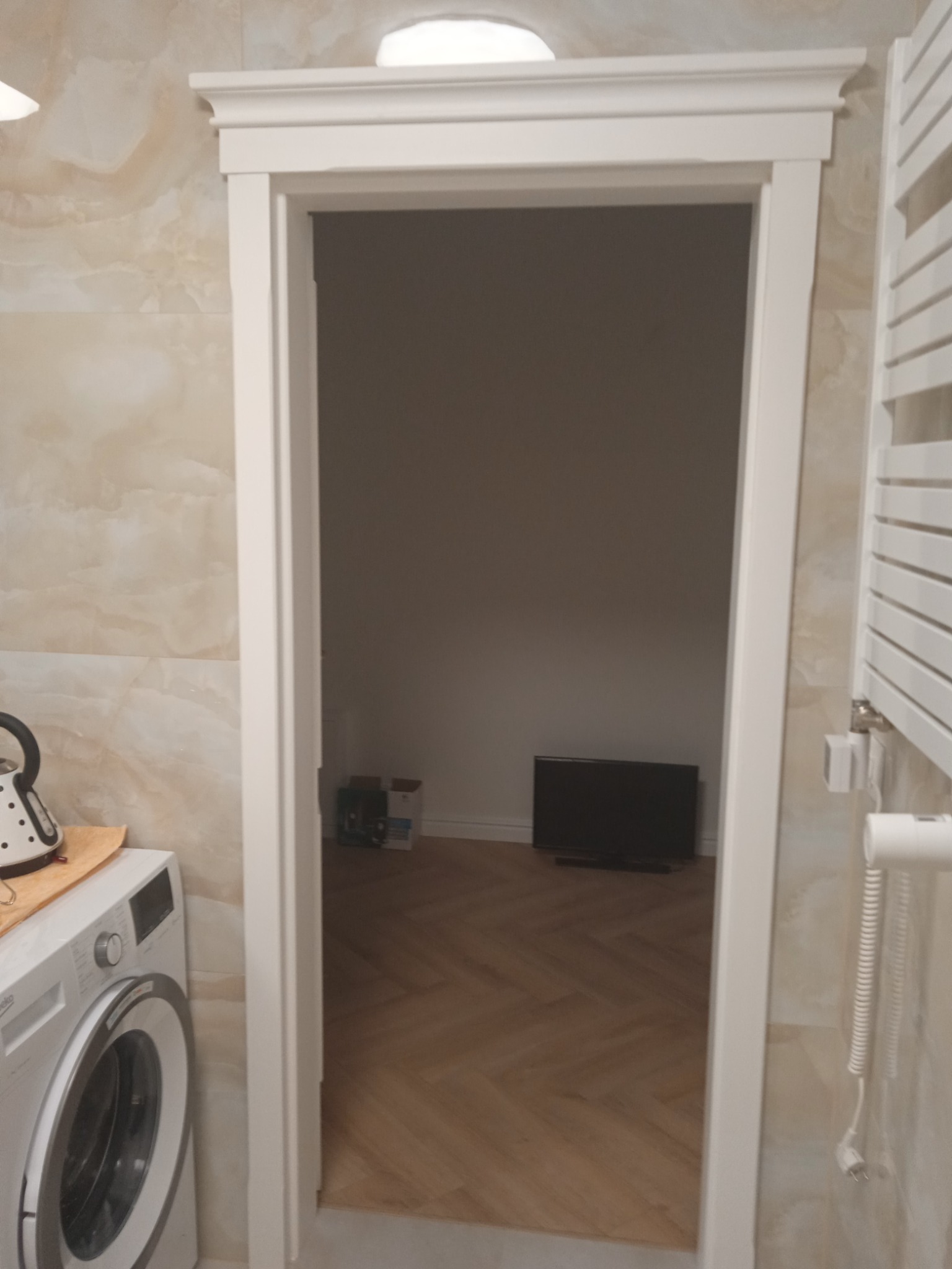 Wykończone przejście między łazienką z pralką i grzejnikiem drabinkowym a pokojem z parkietem w jodełkę i telewizorem, biała listwa wykończeniowa.